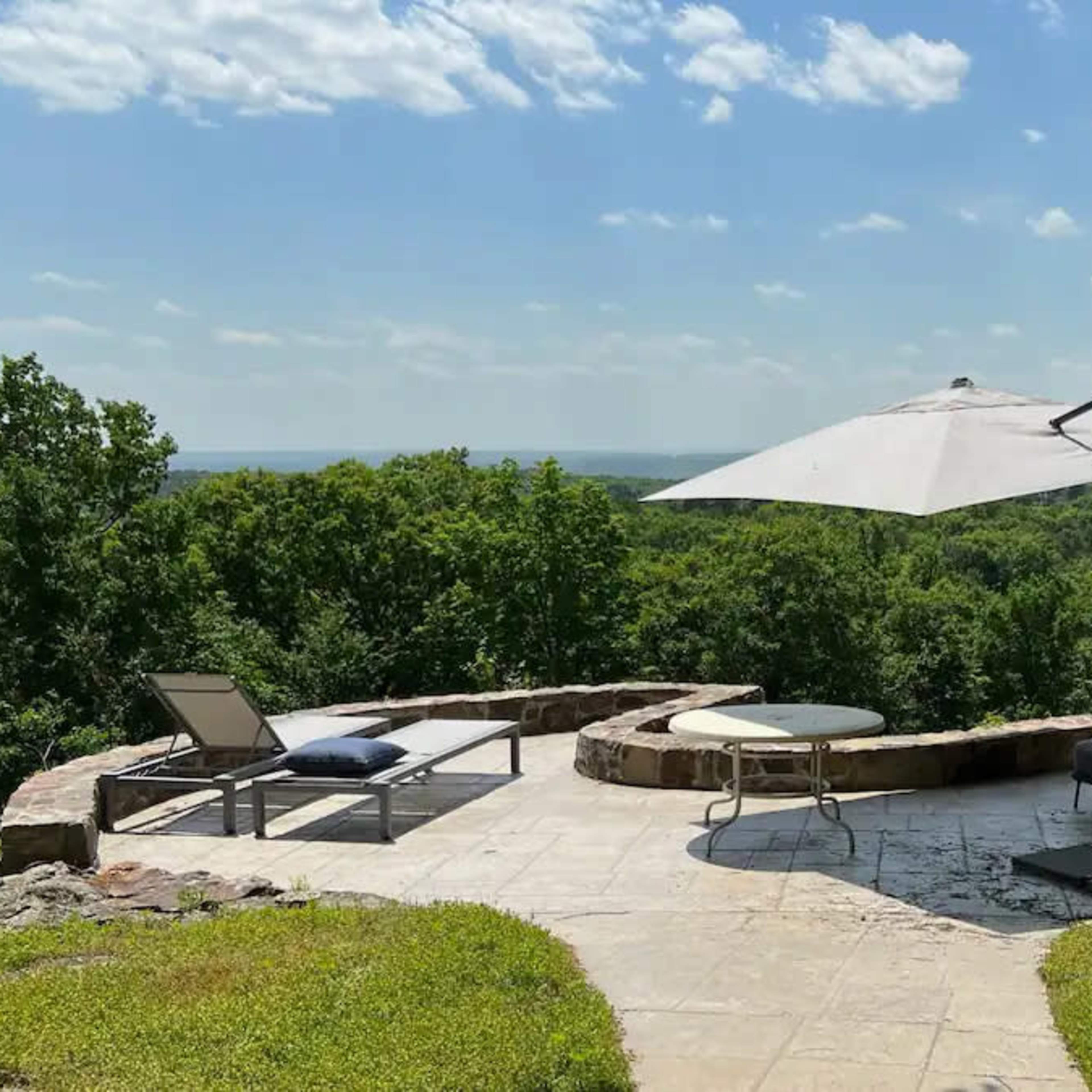 A patio with a seating area, an umbrella, and a view of dense greenery and a distant landscape under a blue sky.