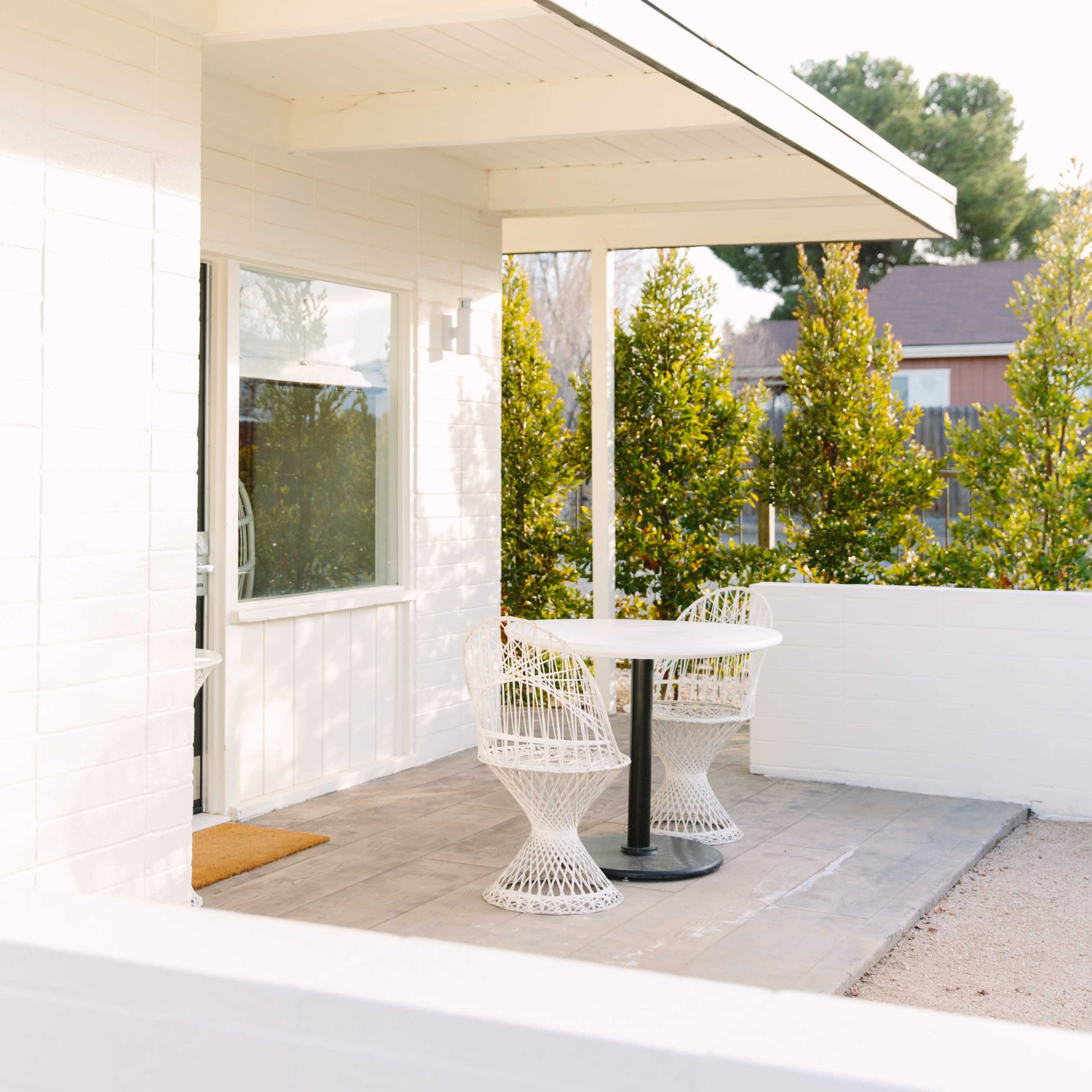 A white patio features a round table surrounded by two woven chairs under a covered area, bordered by greenery.