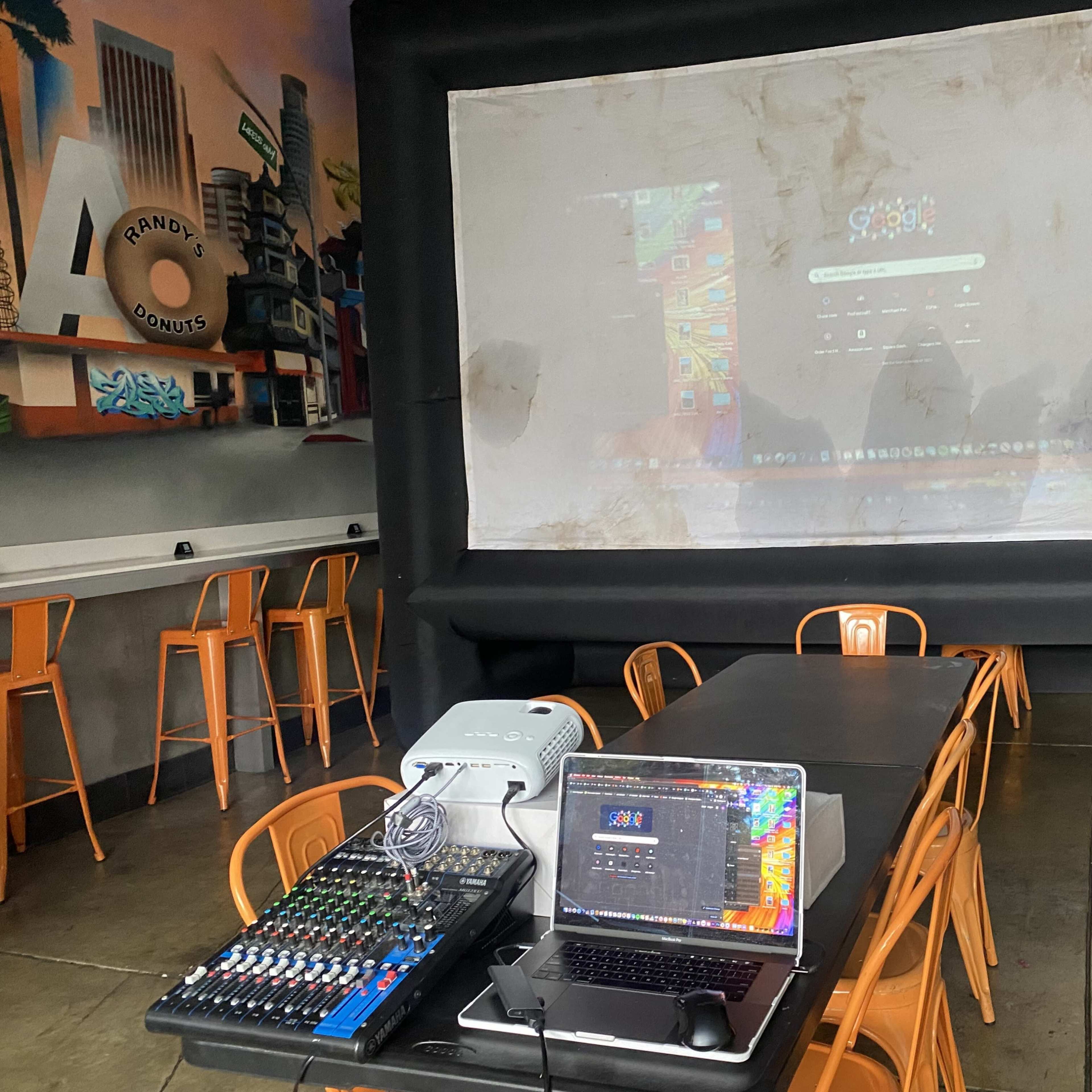 A projector displays a colorful image on a large screen in a dimly lit space with a table and sound equipment set up nearby.