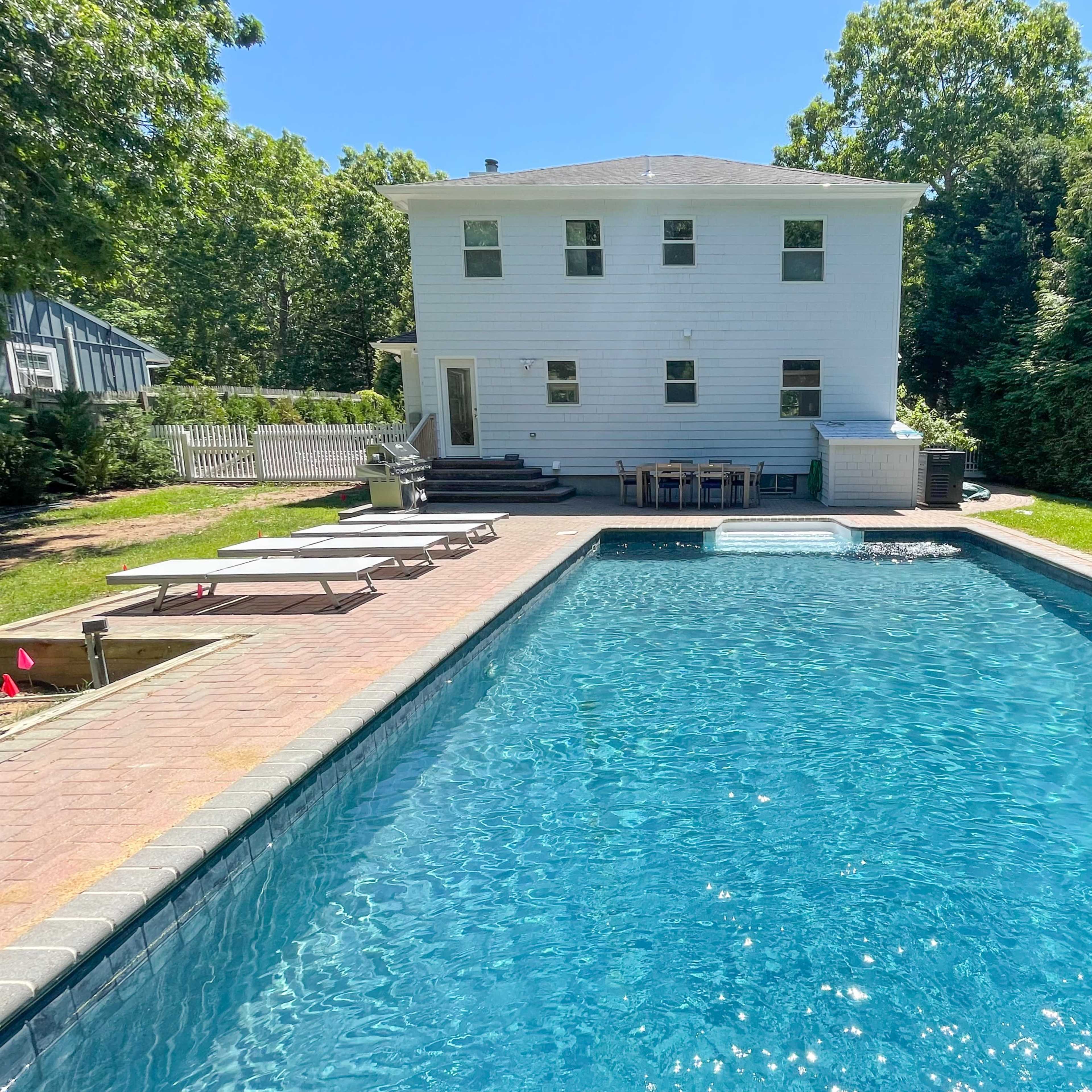 A backyard with a blue swimming pool, surrounded by lounge chairs and a patio area adjacent to a white two-story house.