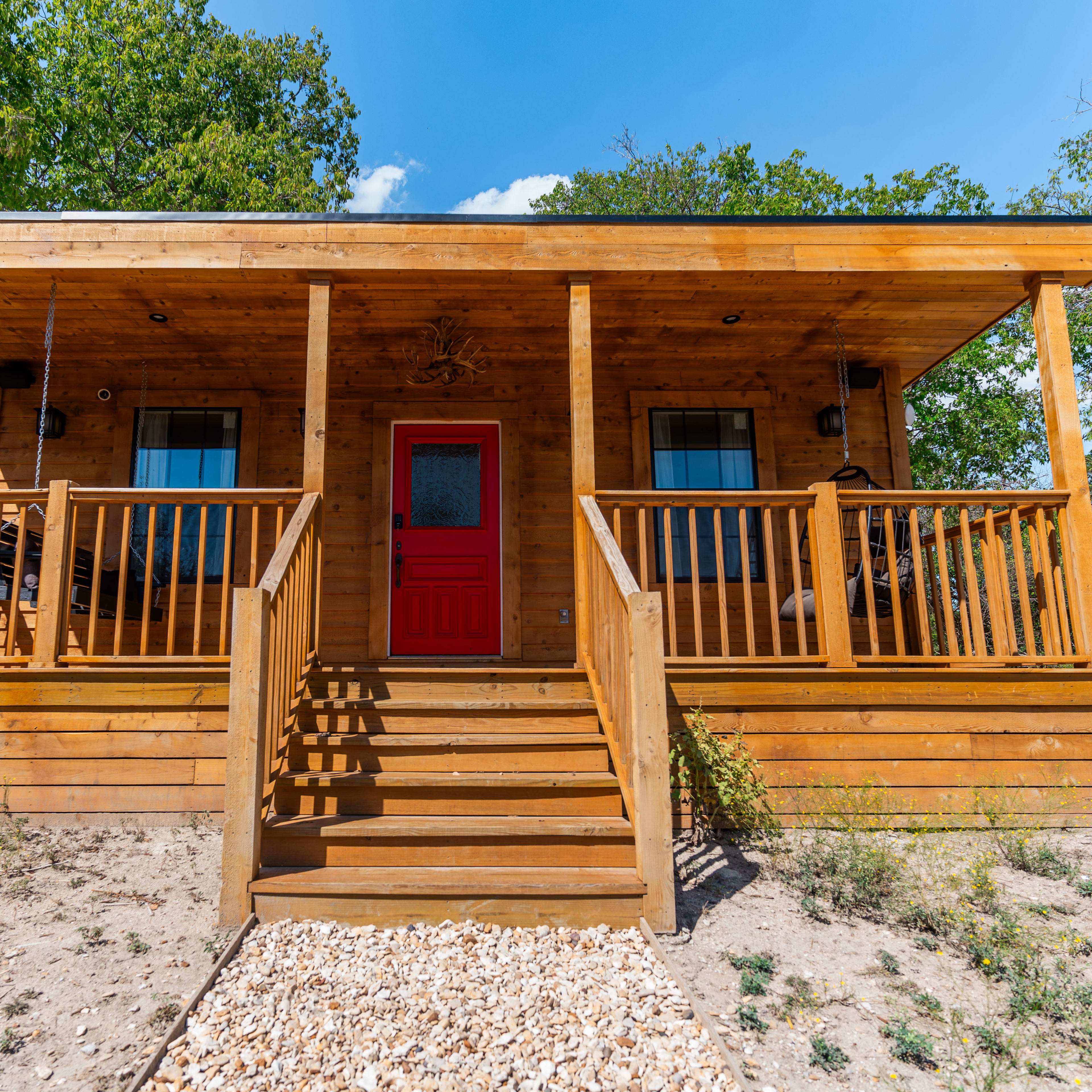A wooden cabin with a red front door and a porch sits on a sandy area, surrounded by trees.
