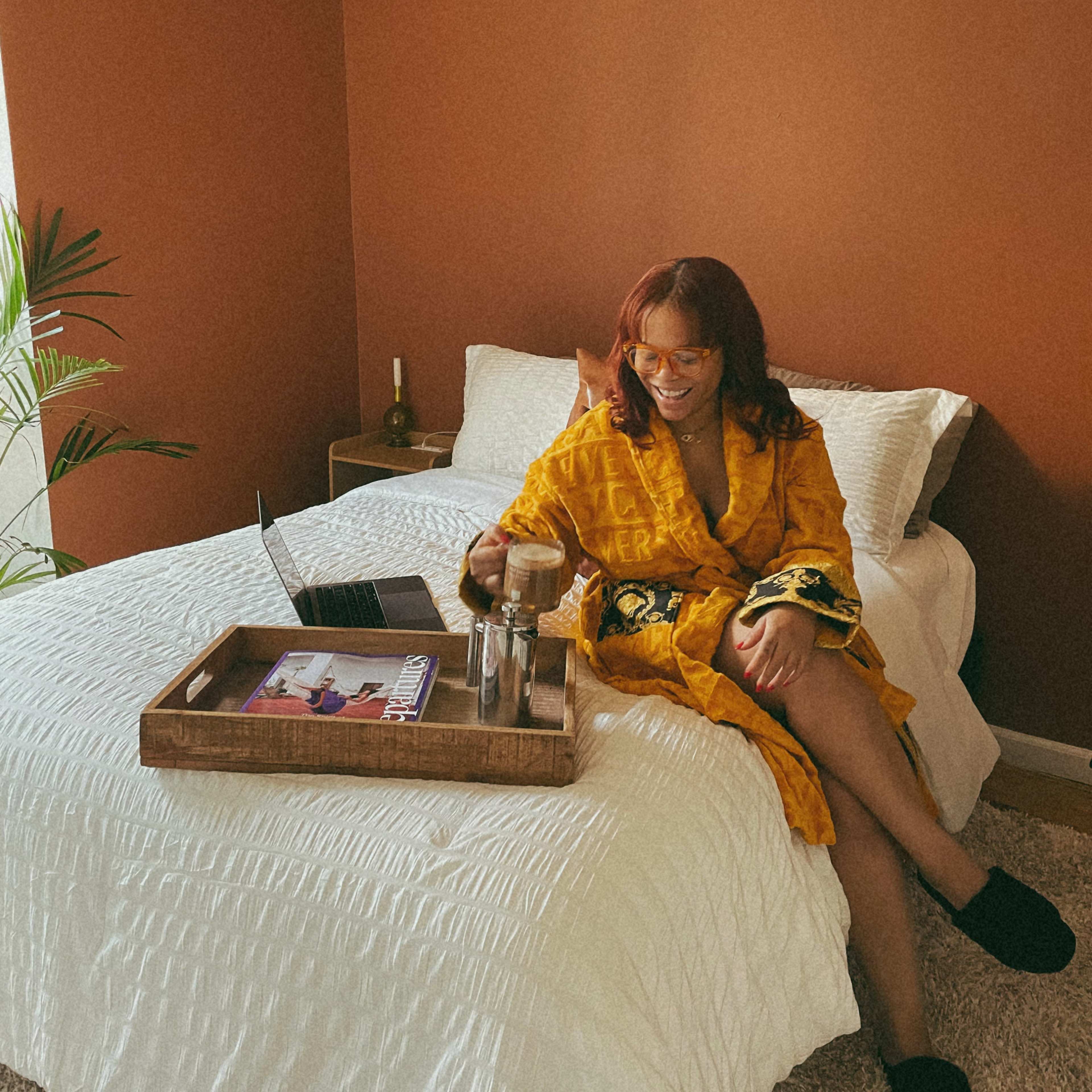 A woman in a yellow robe sits on a bed with a laptop and a tray of drinks in a room with brown walls.