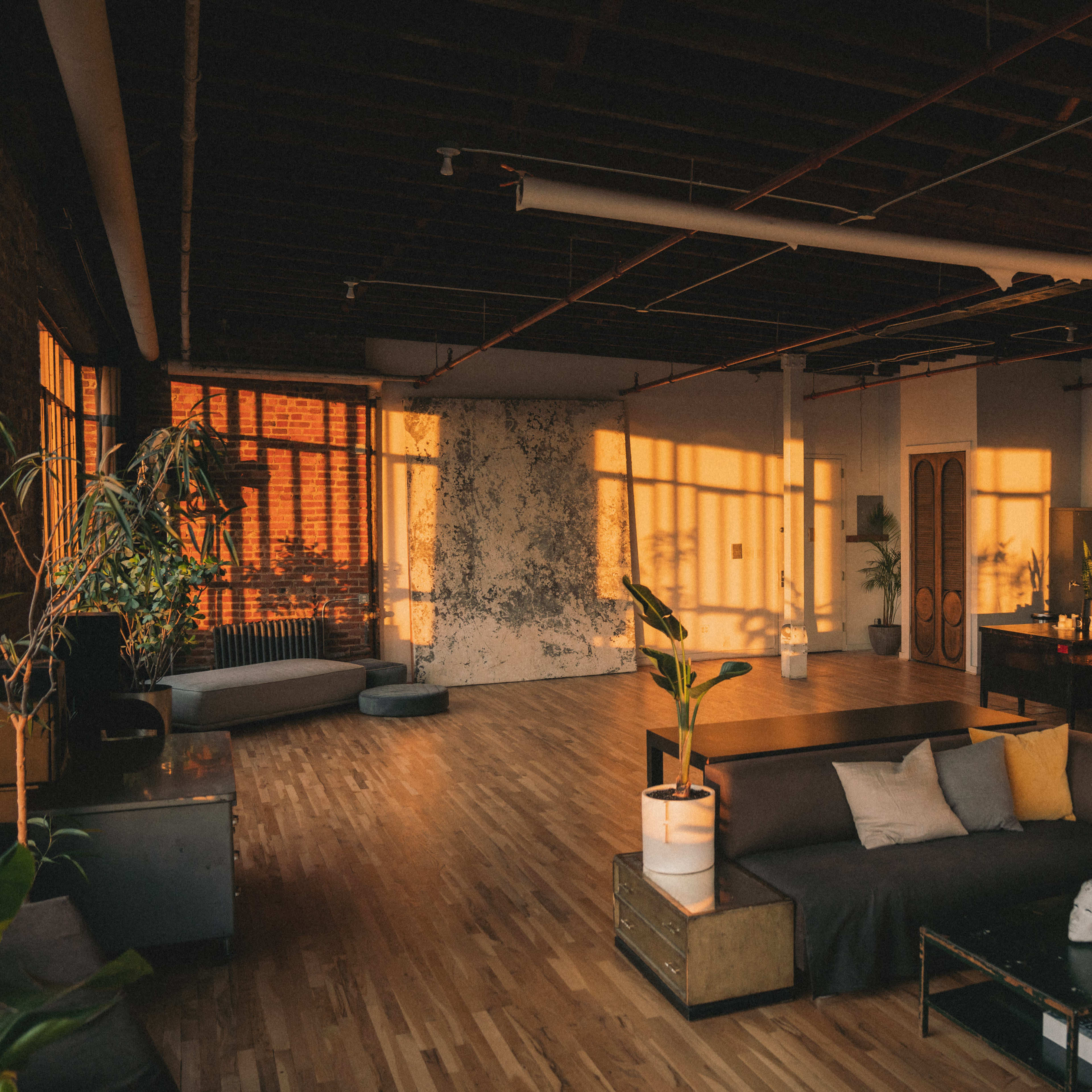 The spacious living area features a large sofa, wooden flooring, and sunlight casting shadows across the walls.
