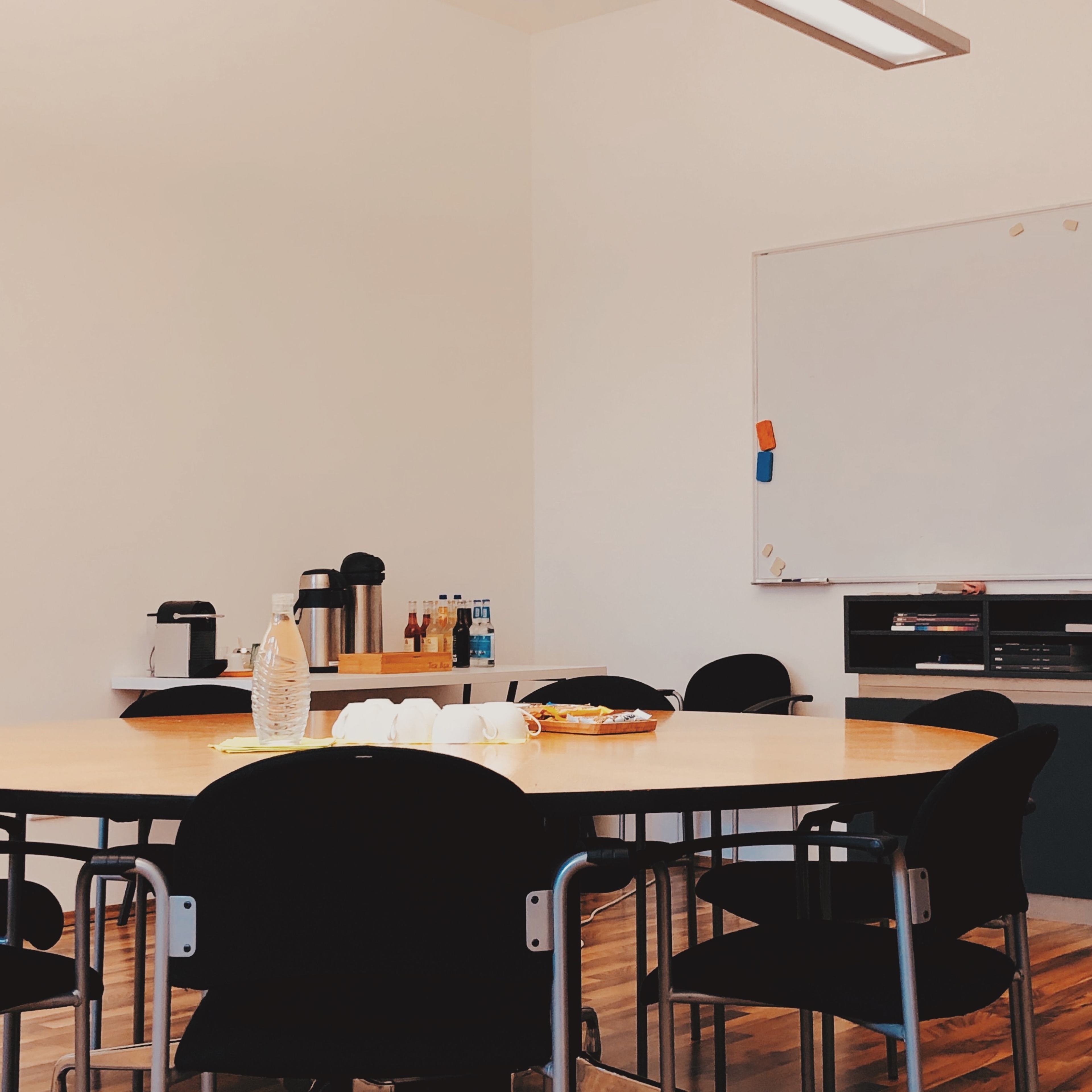 A conference room features a large round table surrounded by black chairs, with a whiteboard and a counter displaying various refreshments in the background.