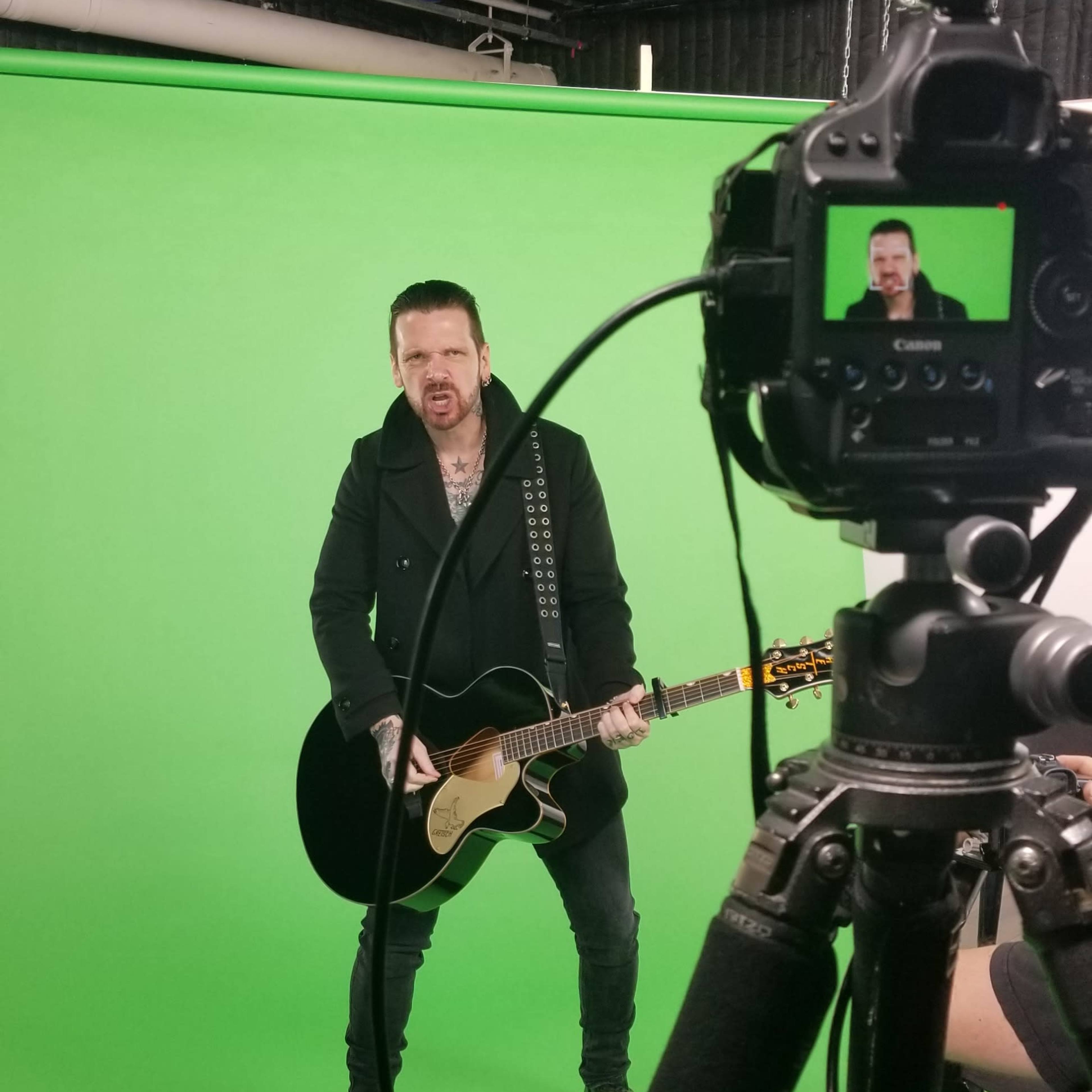 A musician stands with an acoustic guitar in front of a green screen while being filmed by a camera on a tripod.