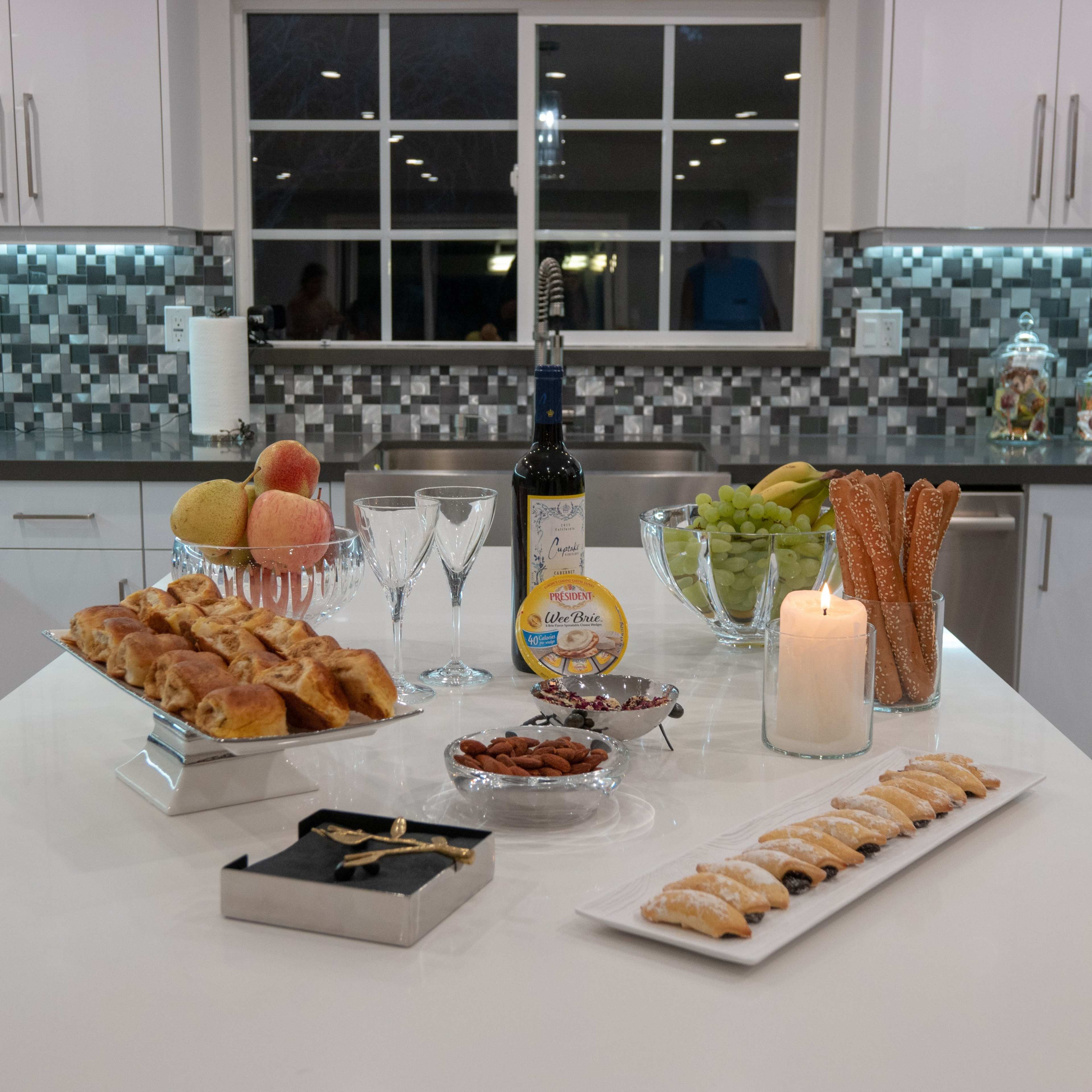 A countertop in a modern kitchen displays an assortment of food items, including pastries, fruits, and snacks, along with a bottle of wine and decorative glasses.