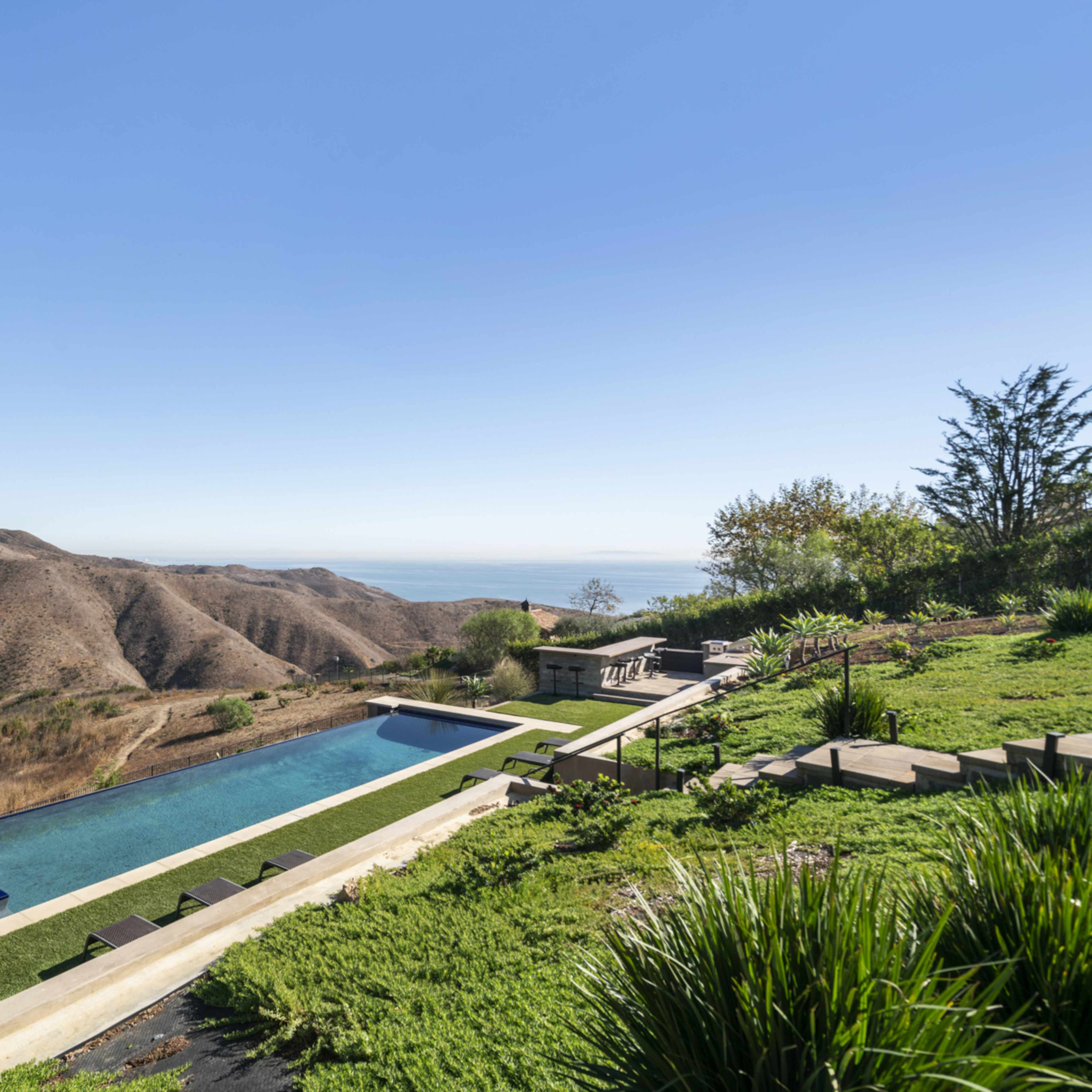 A modern house overlooking a long rectangular swimming pool and rolling hills leading to the ocean in the distance.