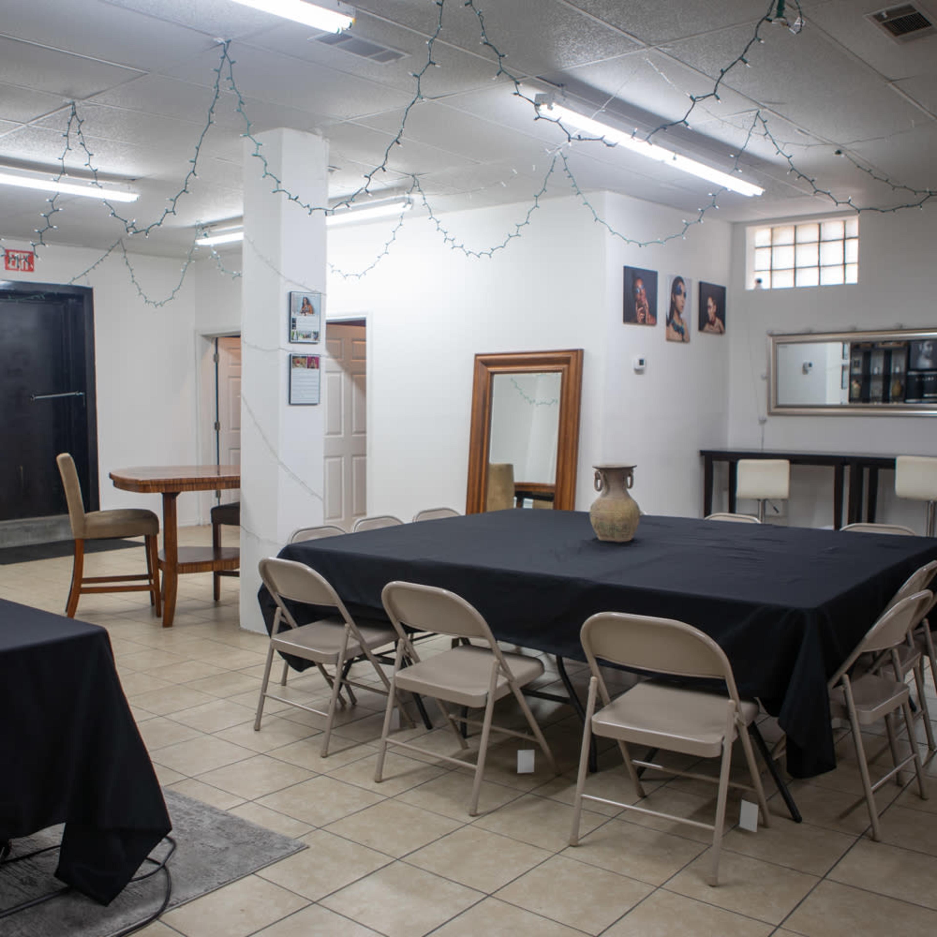A spacious room with a large black table surrounded by folding chairs, with additional seating and string lights hanging from the ceiling.