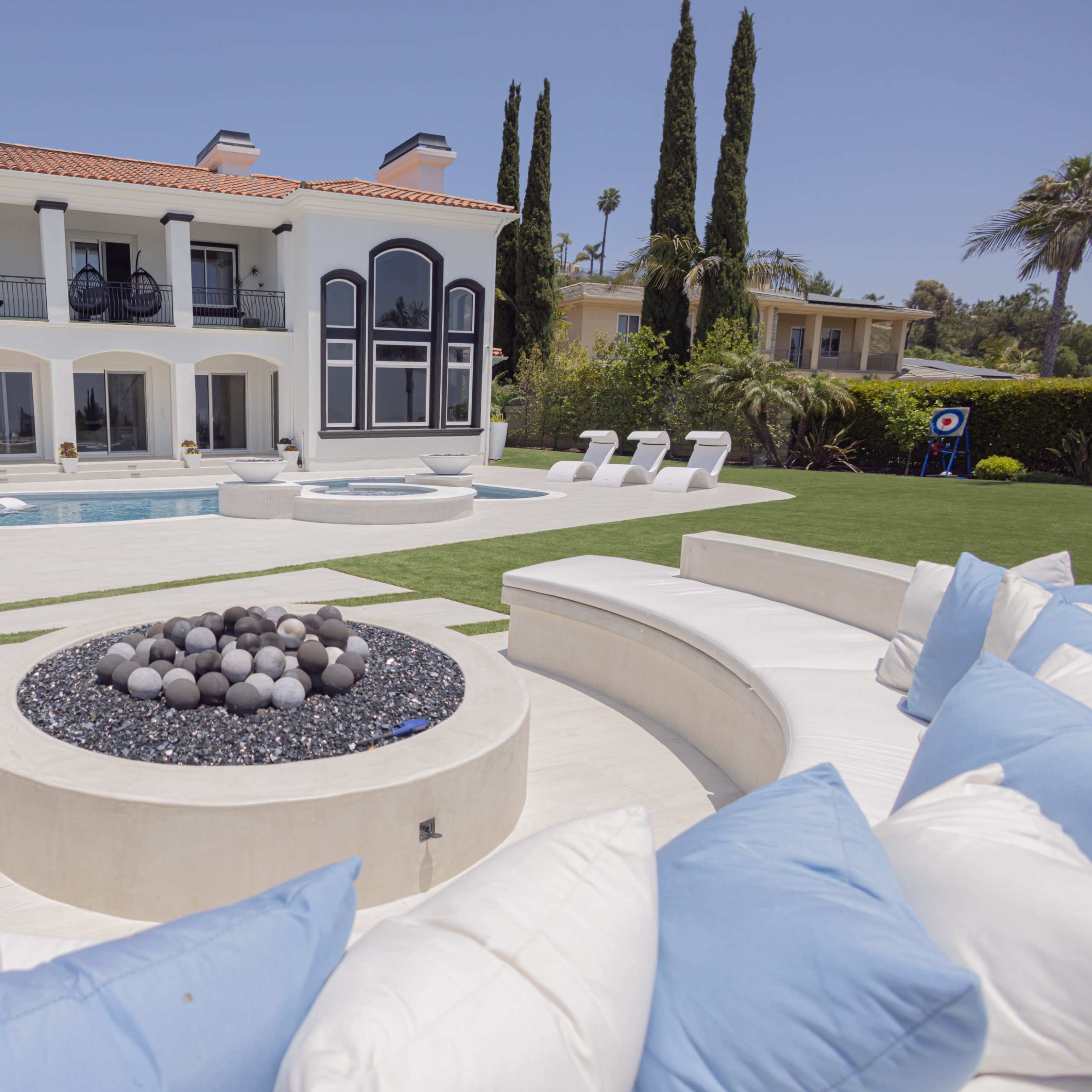 A luxurious outdoor area featuring a modern two-story villa, a swimming pool, and a circular seating area with a fire pit.
