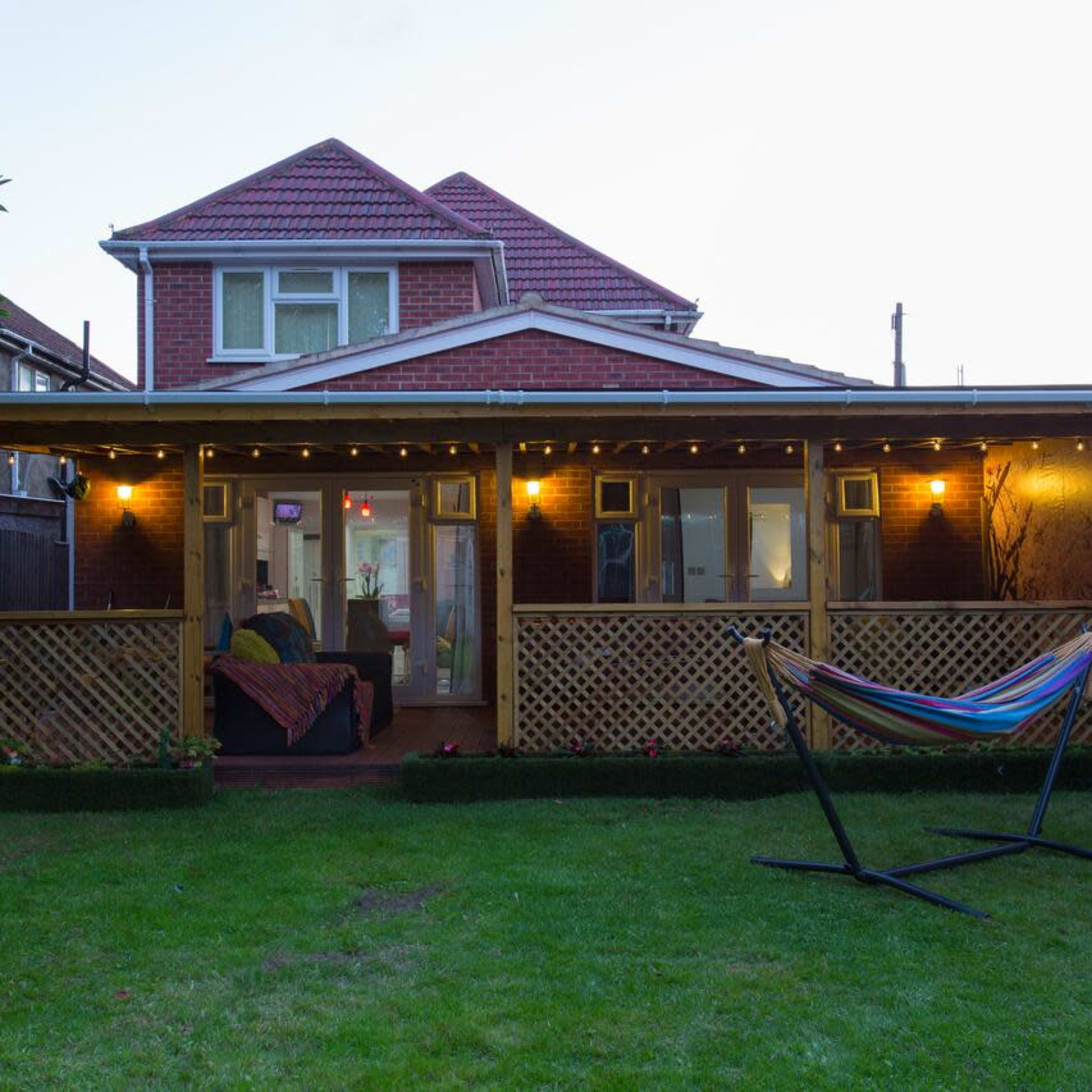 A backyard with a wooden porch illuminated by lights, a hammock stand, and a house in the background.