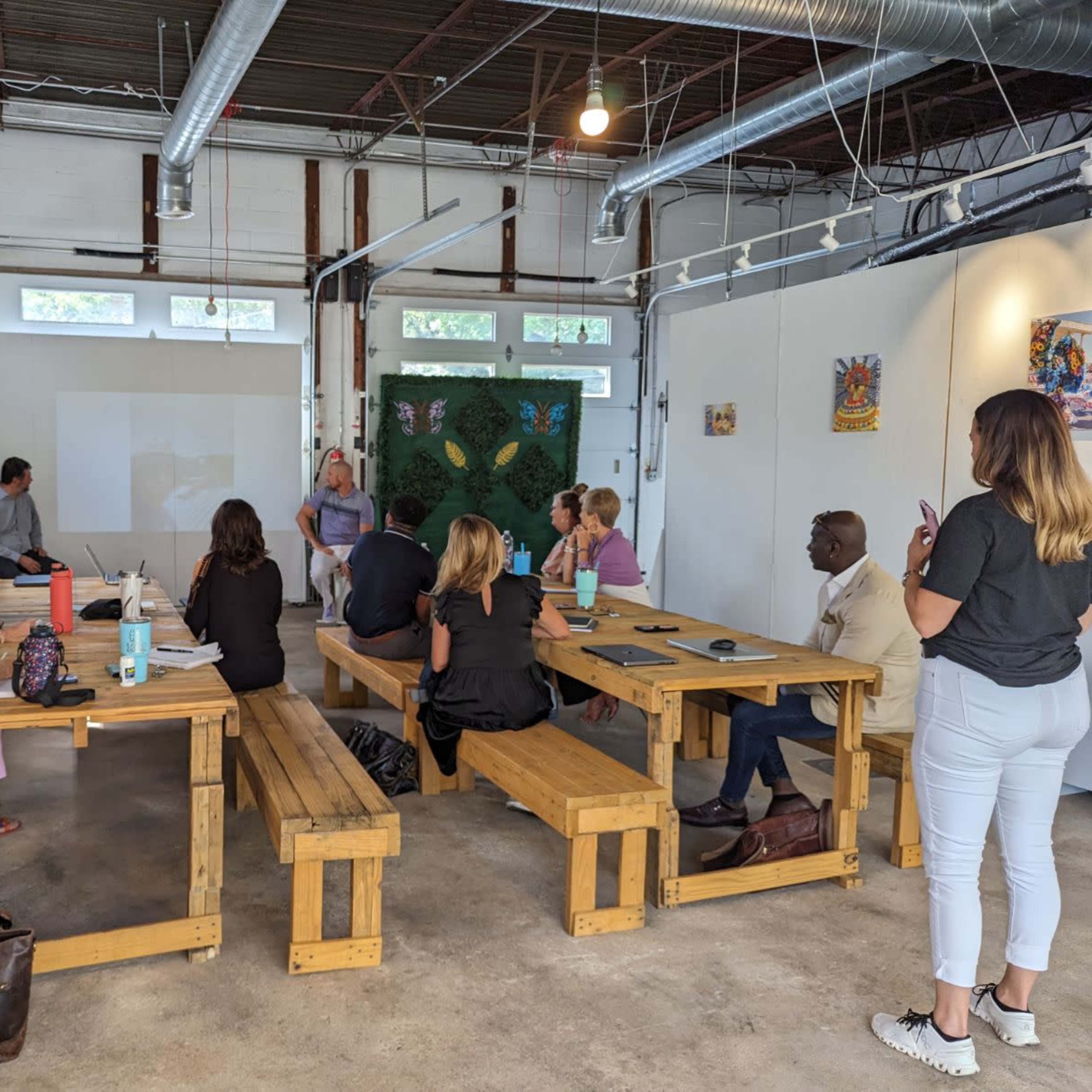 A group of people sits around wooden tables in a bright, industrial-style workspace during a meeting or presentation.