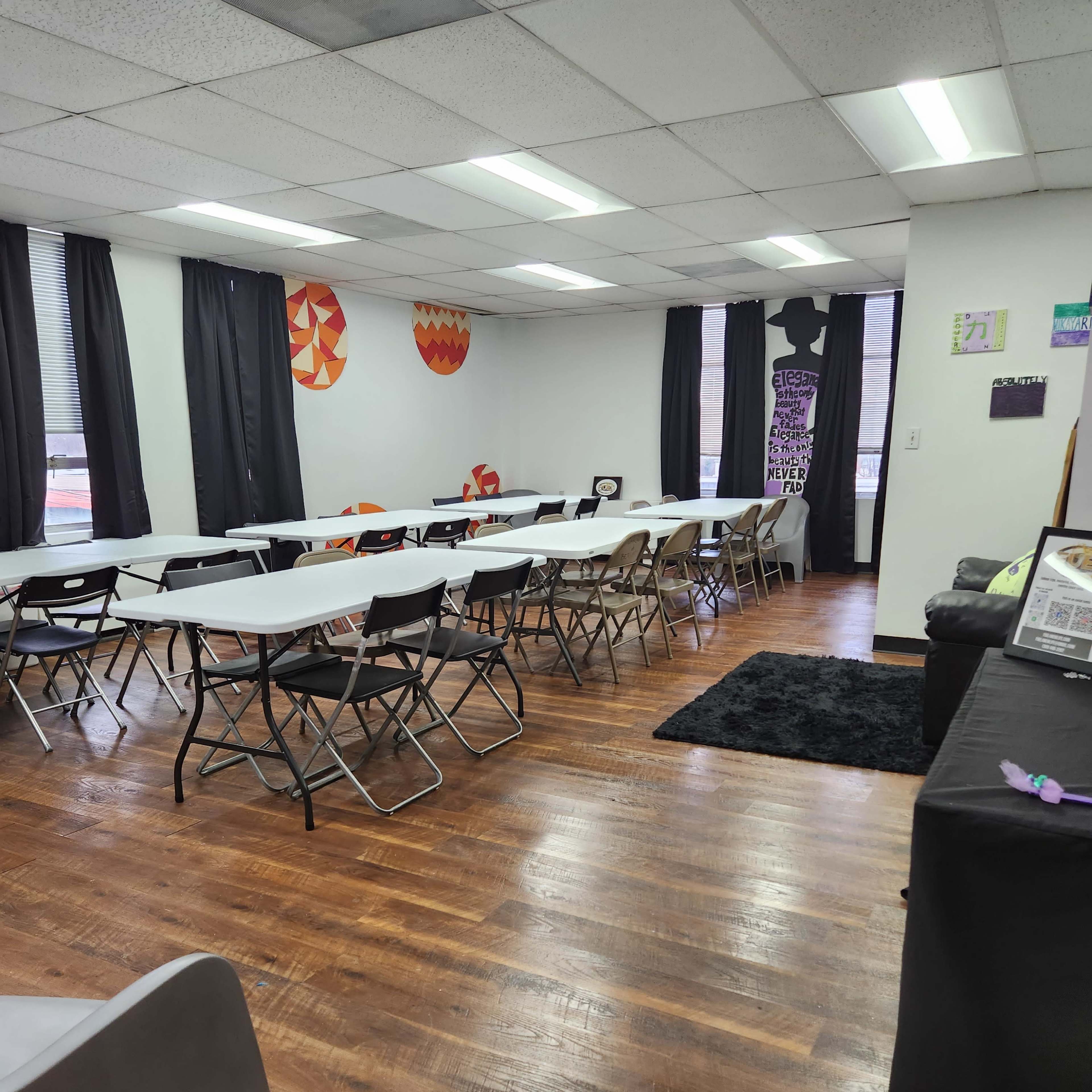 A spacious room set up with several folding tables and chairs arranged for a gathering, with black curtains and colorful wall decorations.
