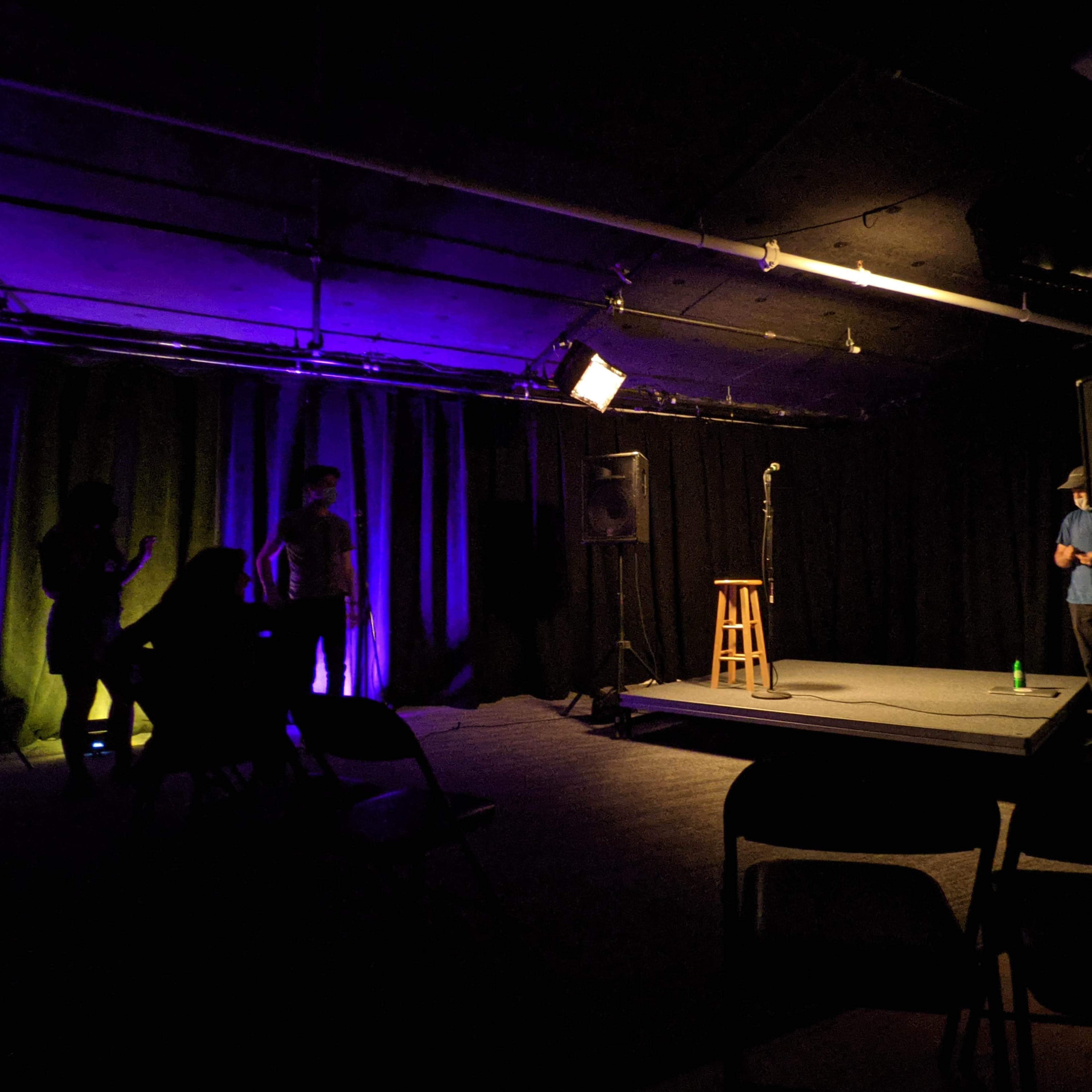 A dimly lit stage with a microphone on a stool, surrounded by empty chairs and silhouettes of people in the background.
