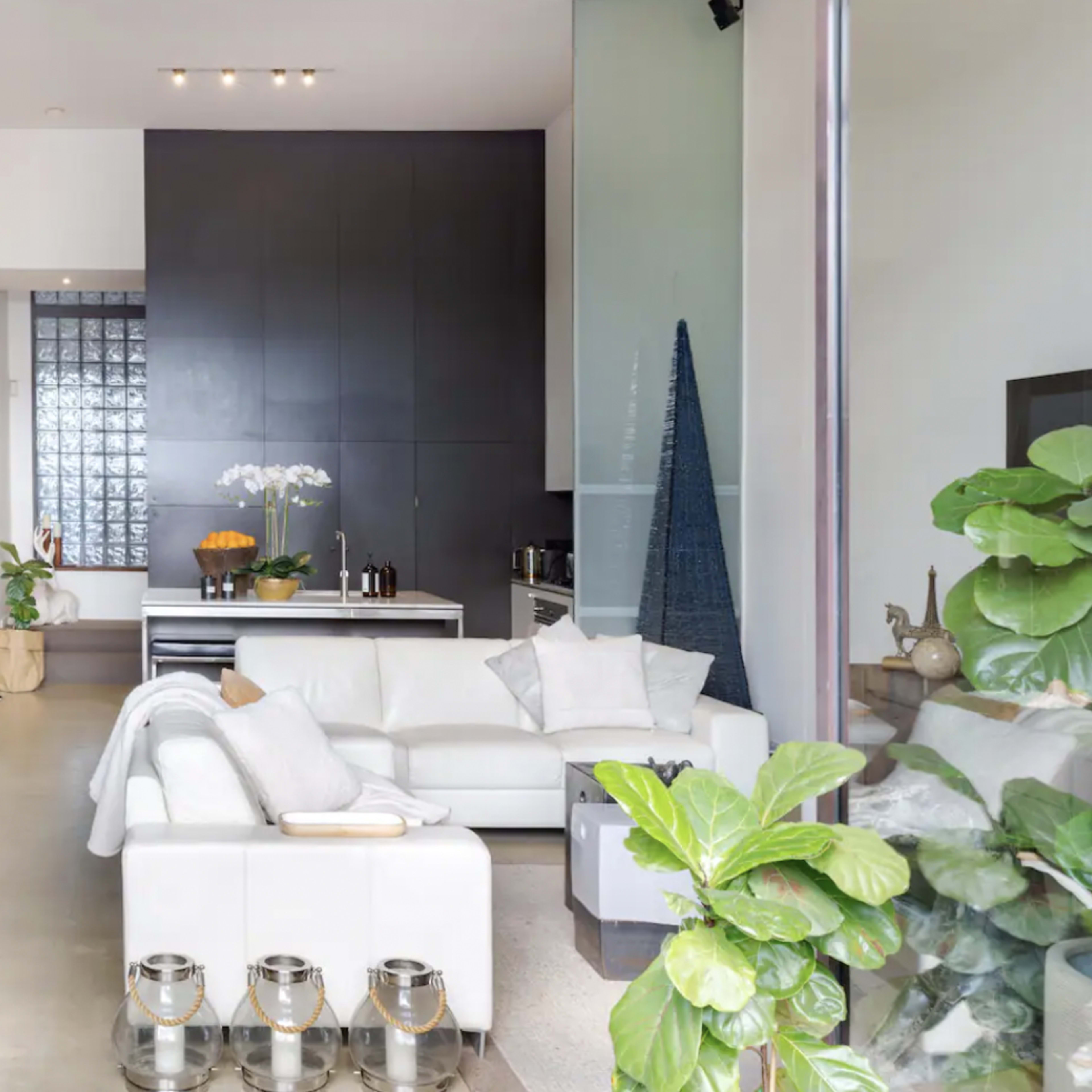 A modern living space featuring a white sectional sofa, a dining area with woven chairs, and large plants, set against a backdrop of sleek furniture and a glass block wall.