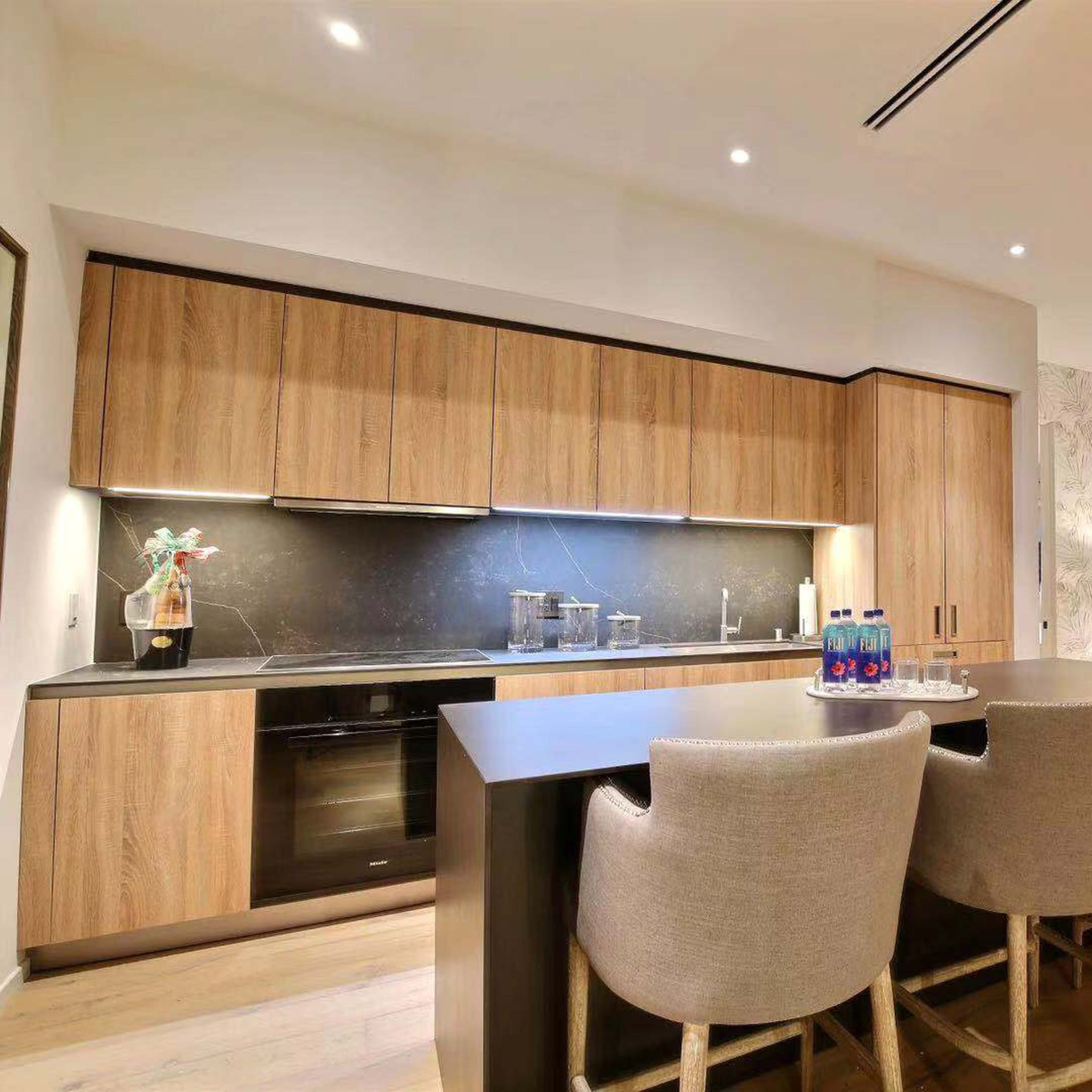 A modern kitchen featuring wooden cabinetry, a large island with seating, and a living area visible in the background.