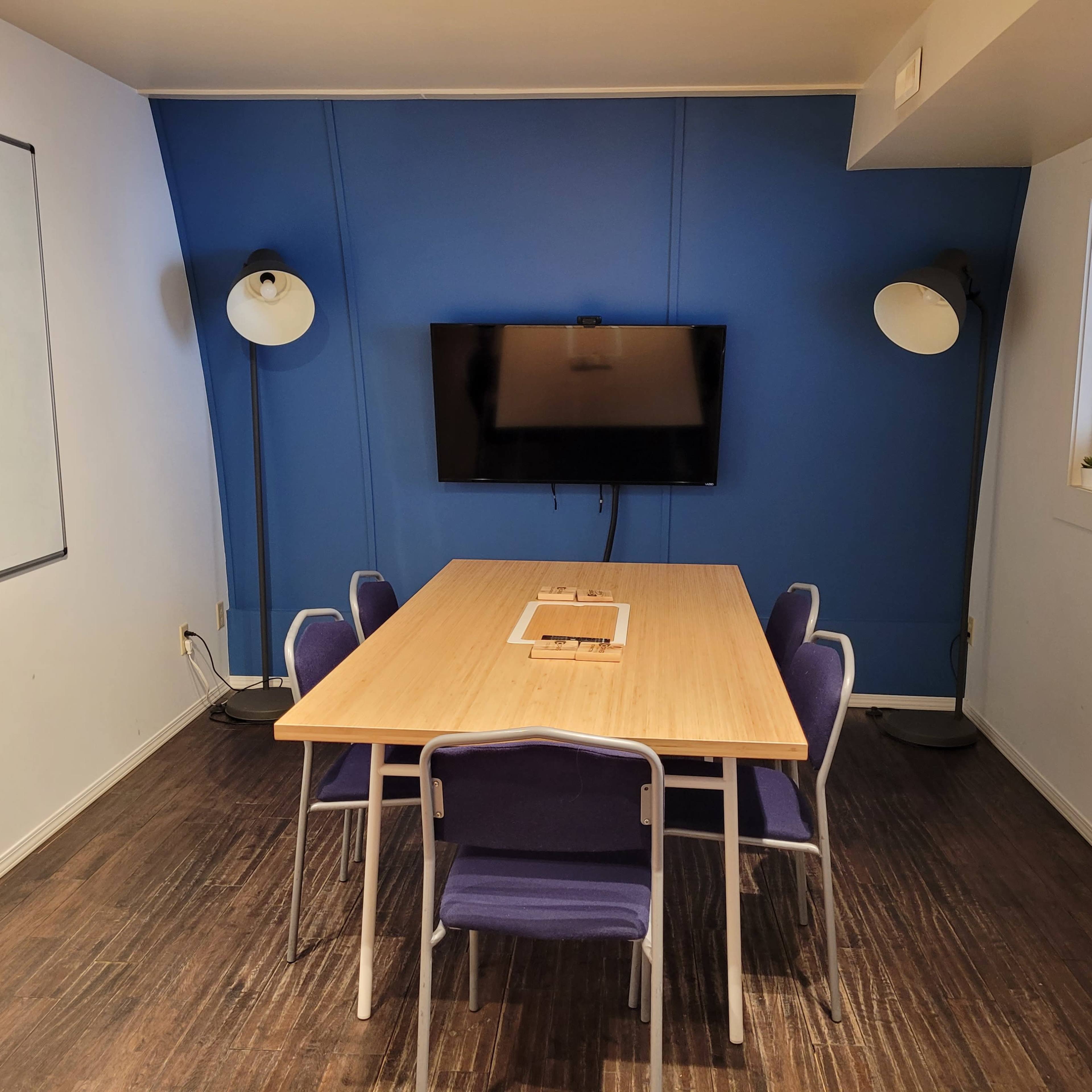 A conference room features a wooden table surrounded by purple chairs, with a blue wall and a television mounted on it.