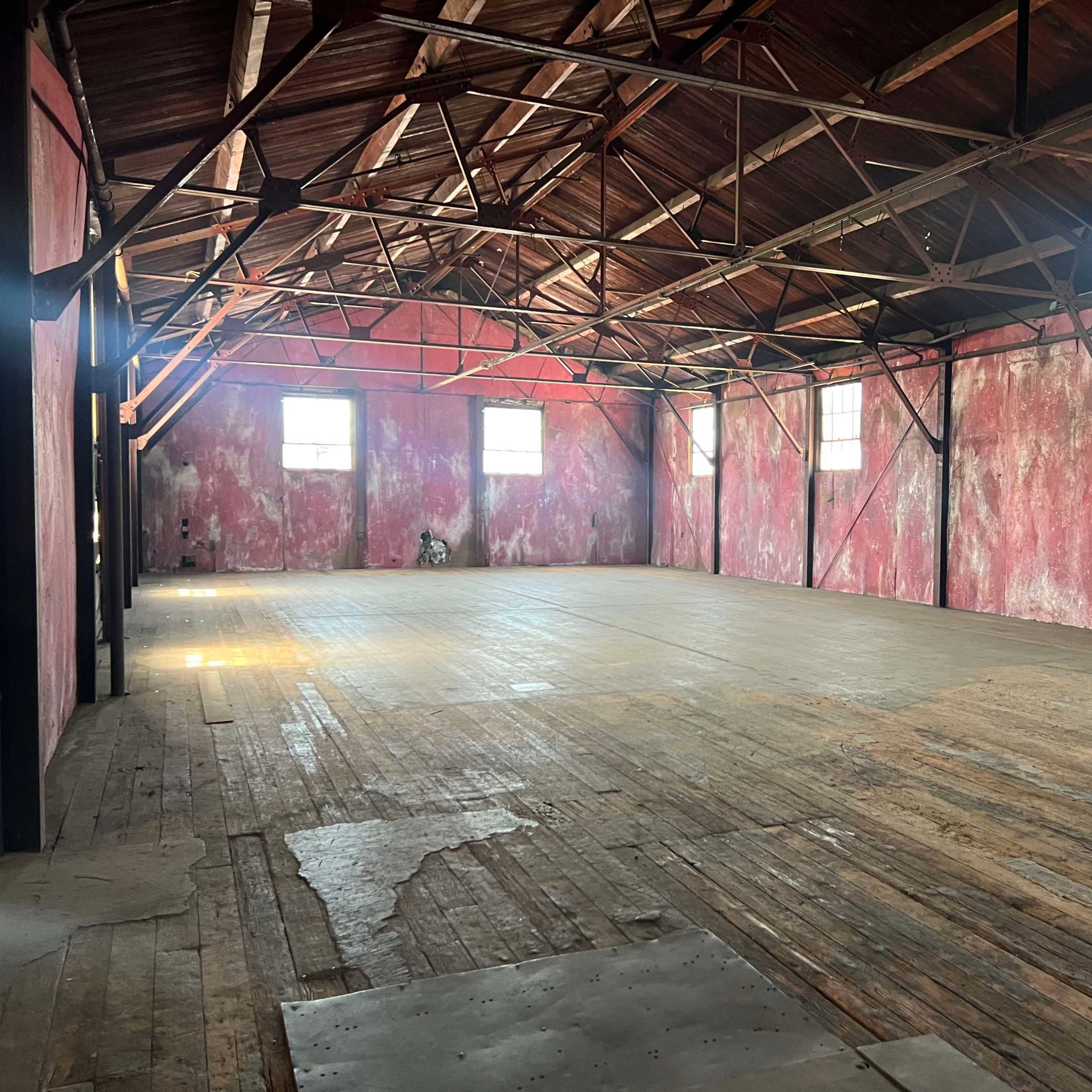 An empty room with wooden flooring, exposed beams, and peeling red paint on the walls, illuminated by natural light coming through the windows.