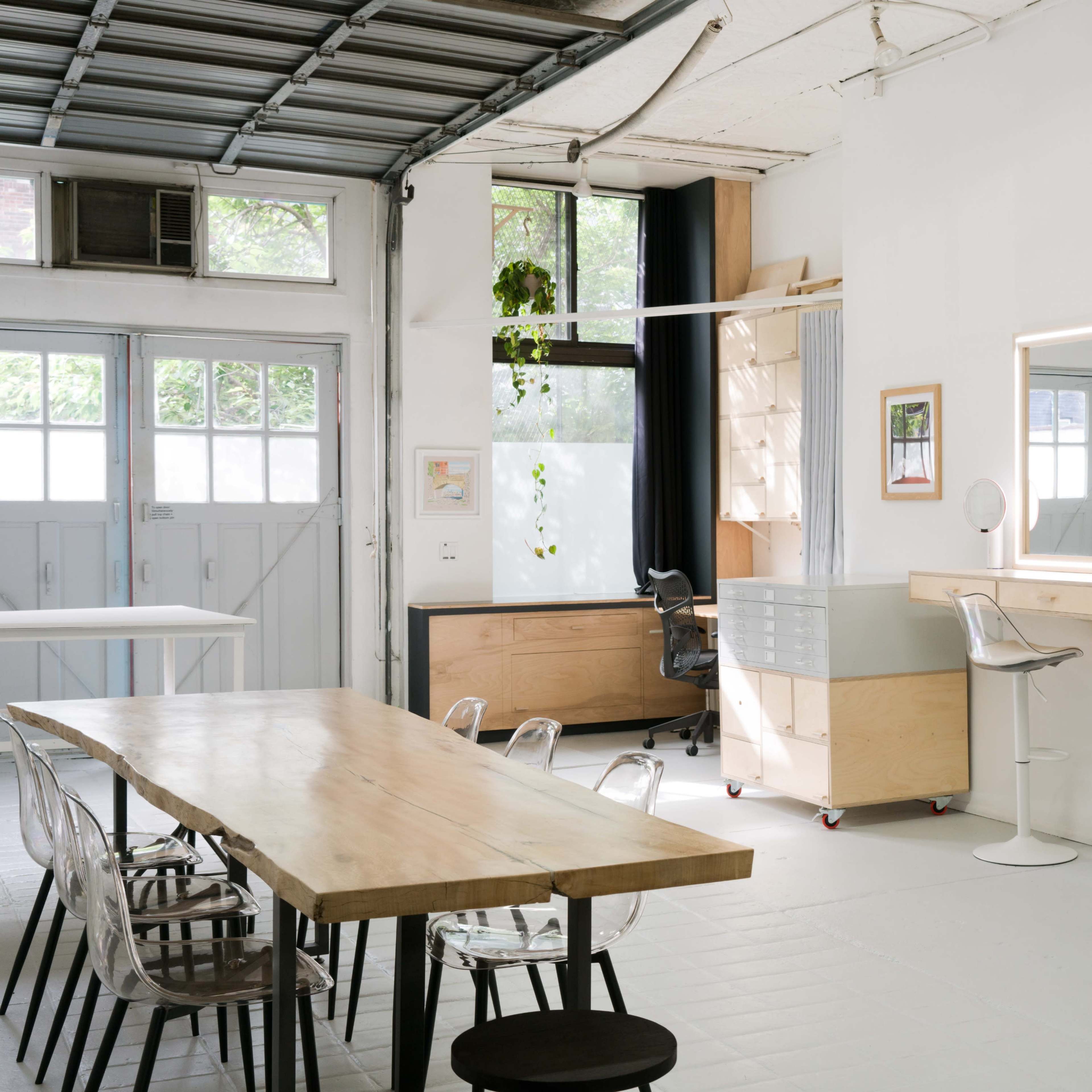 A modern workspace with a large wooden table, clear acrylic chairs, a sleek workspace area with two mirrors, and a garage-style door allowing natural light.