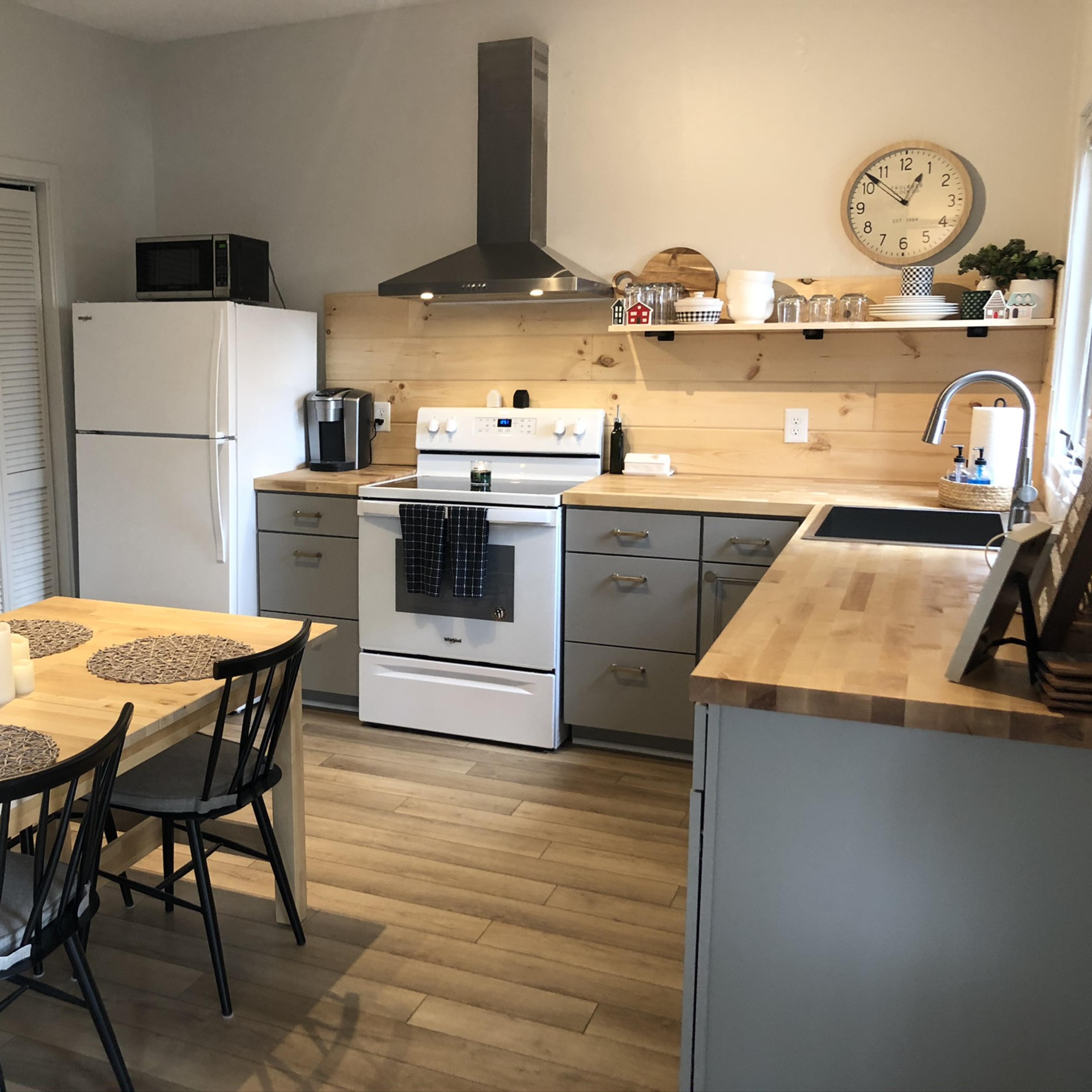 A modern kitchen with a dining table and six chairs, a white stove, a refrigerator, and wooden shelves displaying various kitchen items.