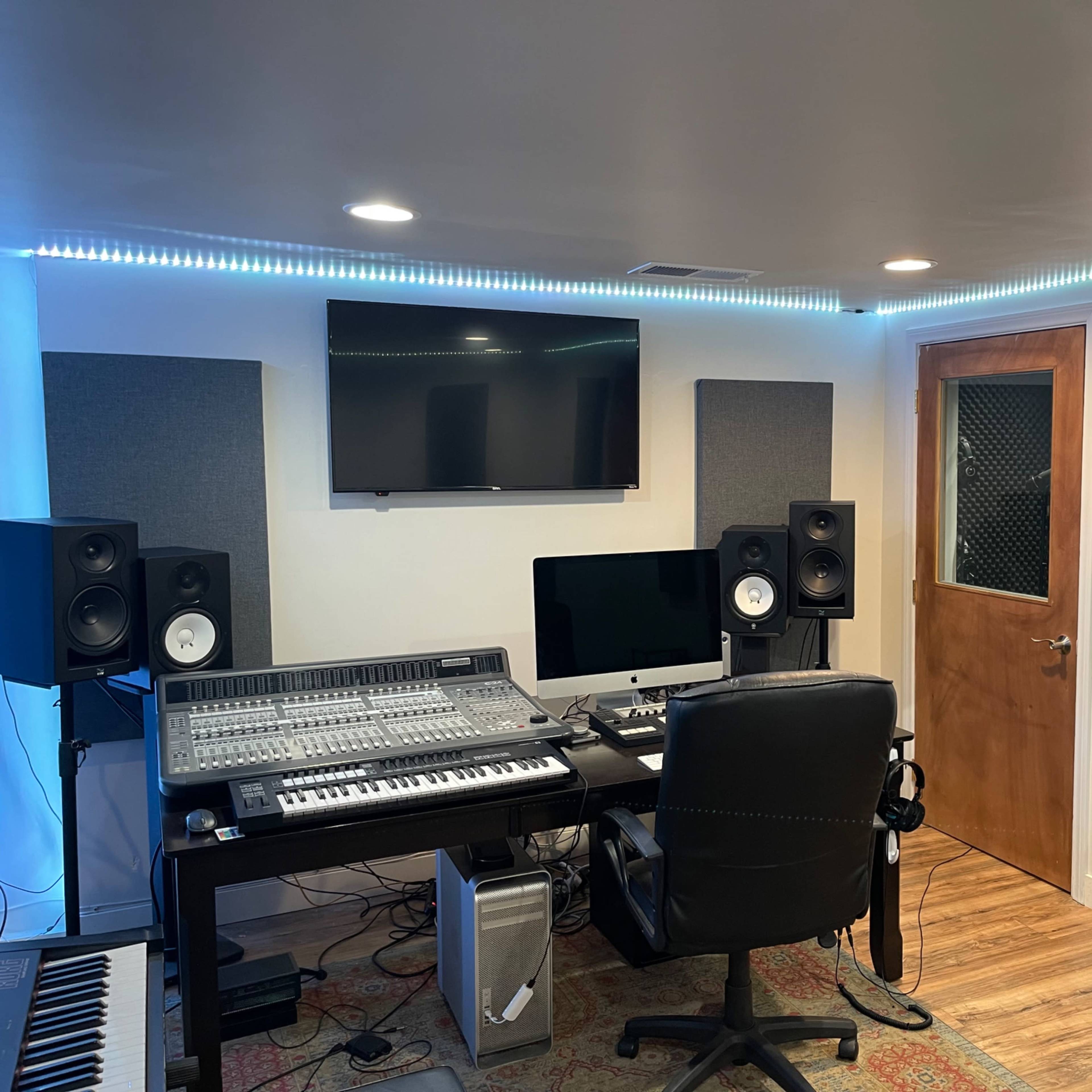 A modern music studio featuring a mixing console, two speakers, a computer, and soundproofing panels, with a chair and door in the background.