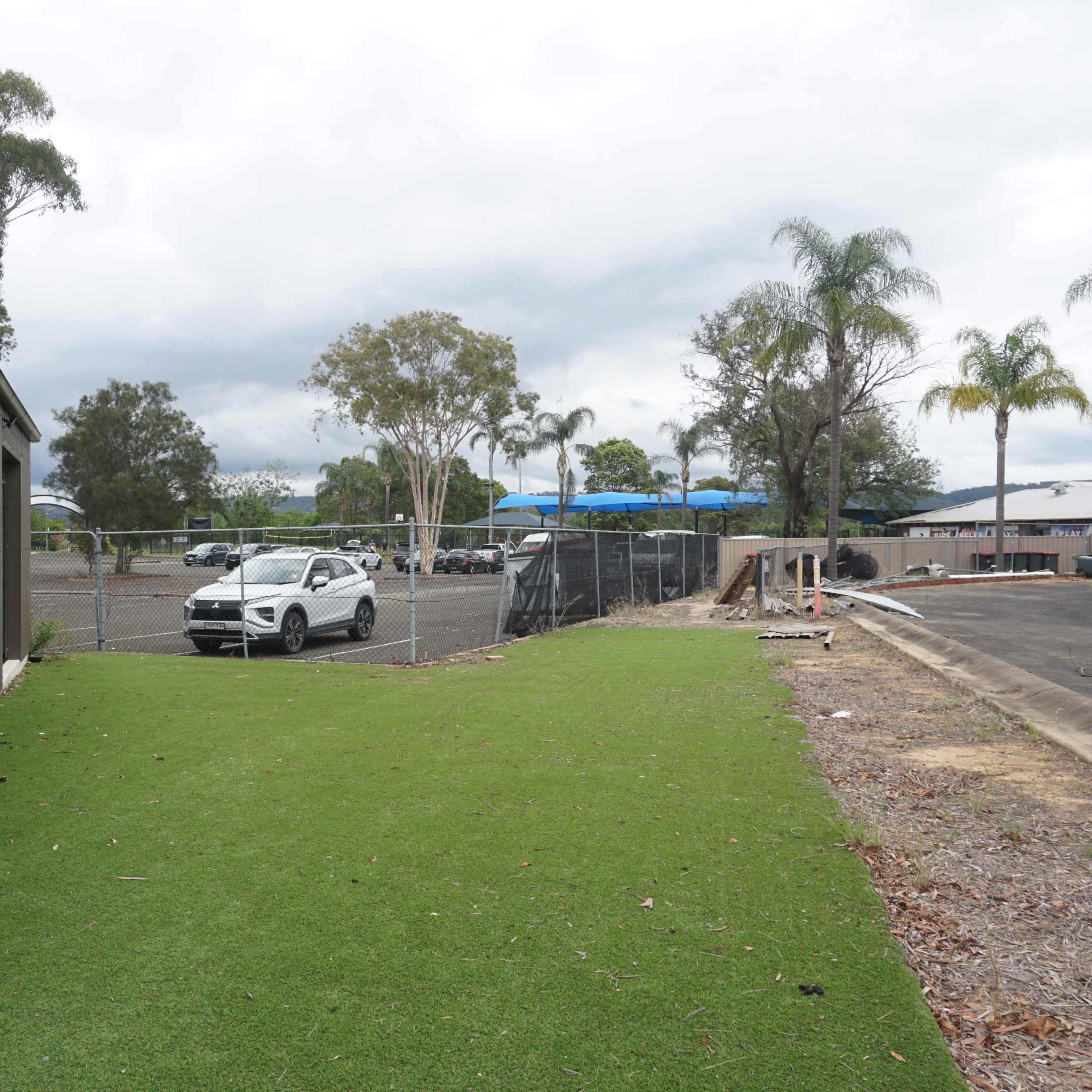 A landscaped area with artificial grass next to a metal building, with a parked car and a fenced lot in the background.