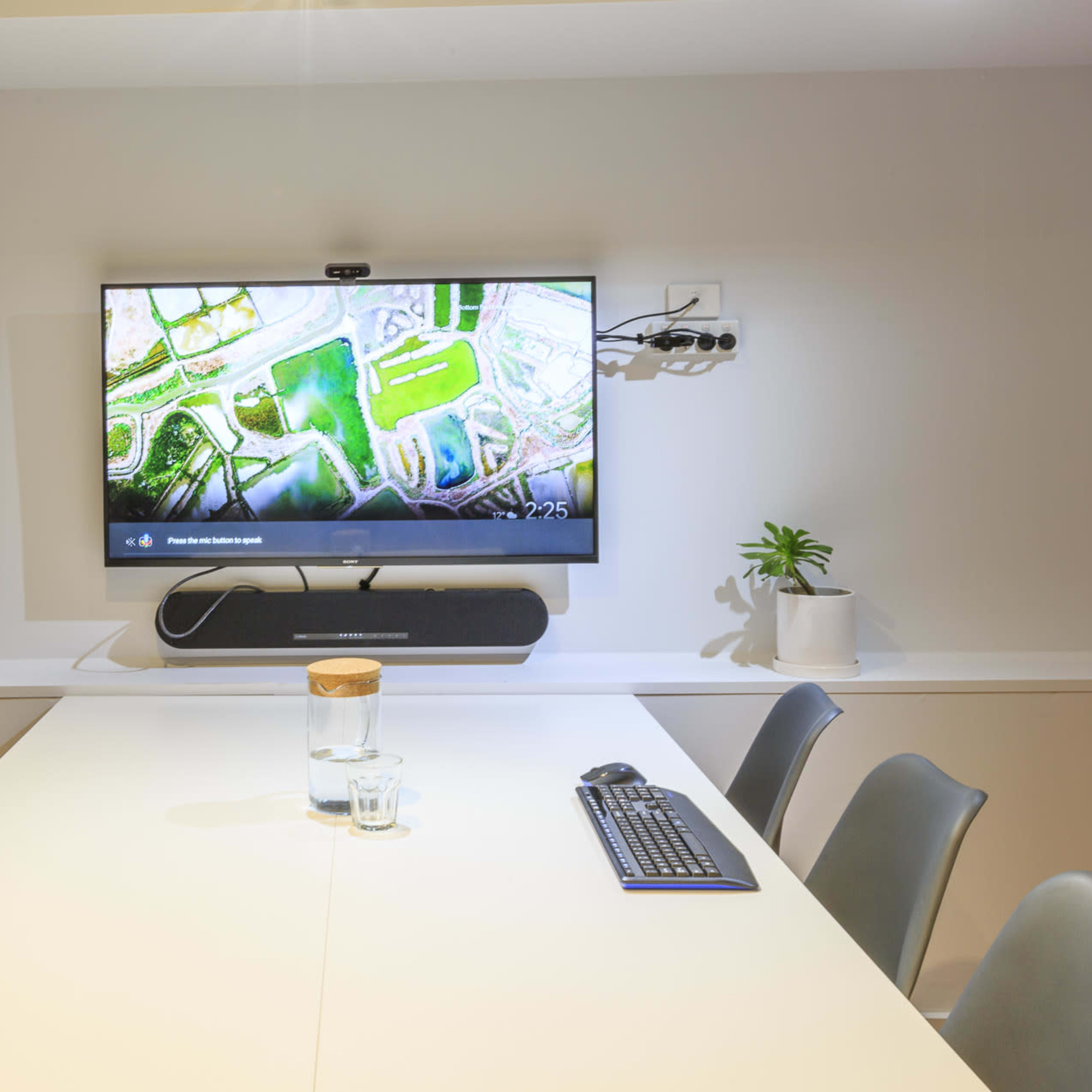 A modern conference room features a large screen displaying a map, with a long table, chairs, a keyboard, a glass of water, and potted plants visible.