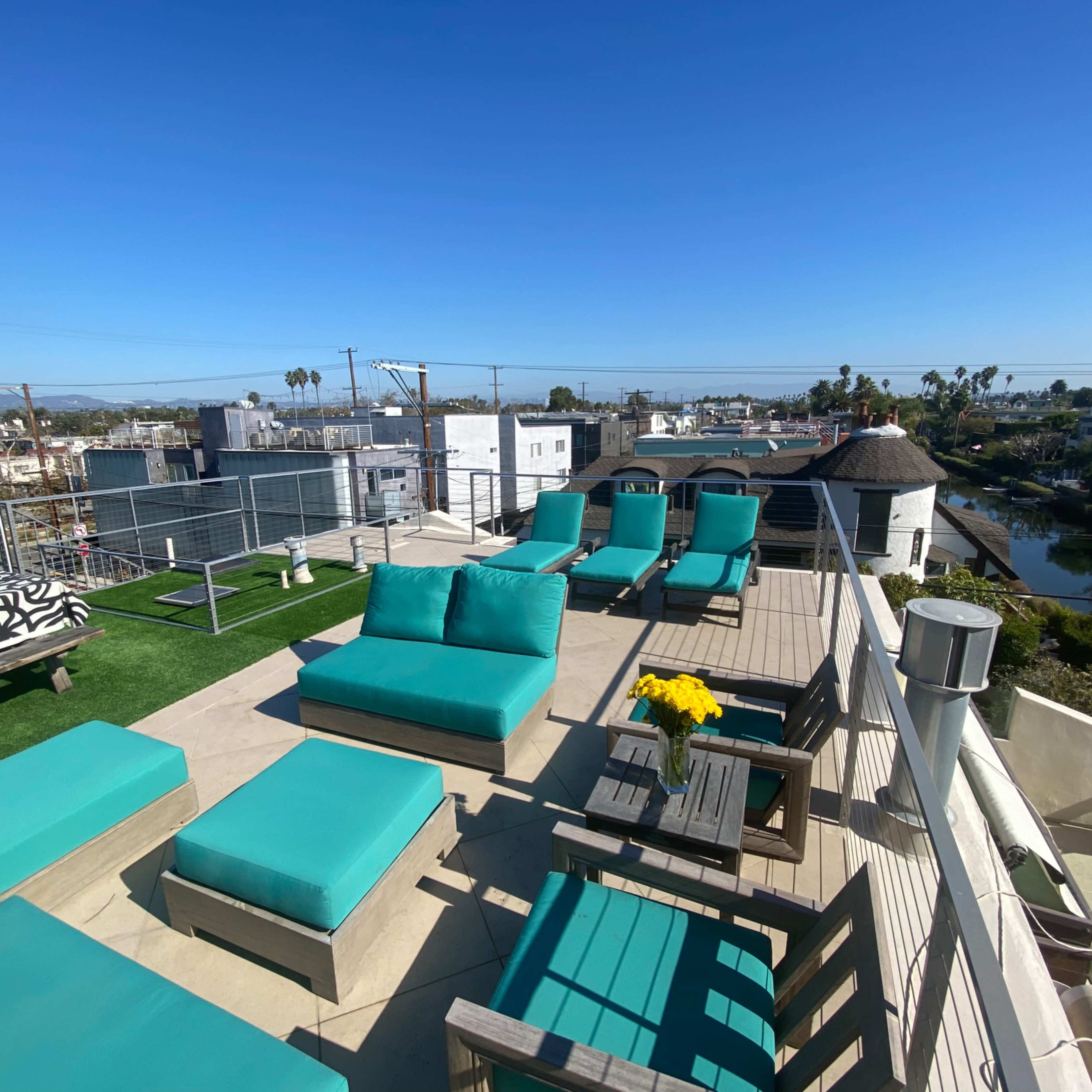 A rooftop terrace features several turquoise lounge chairs and tables with a view of nearby buildings and palm trees.