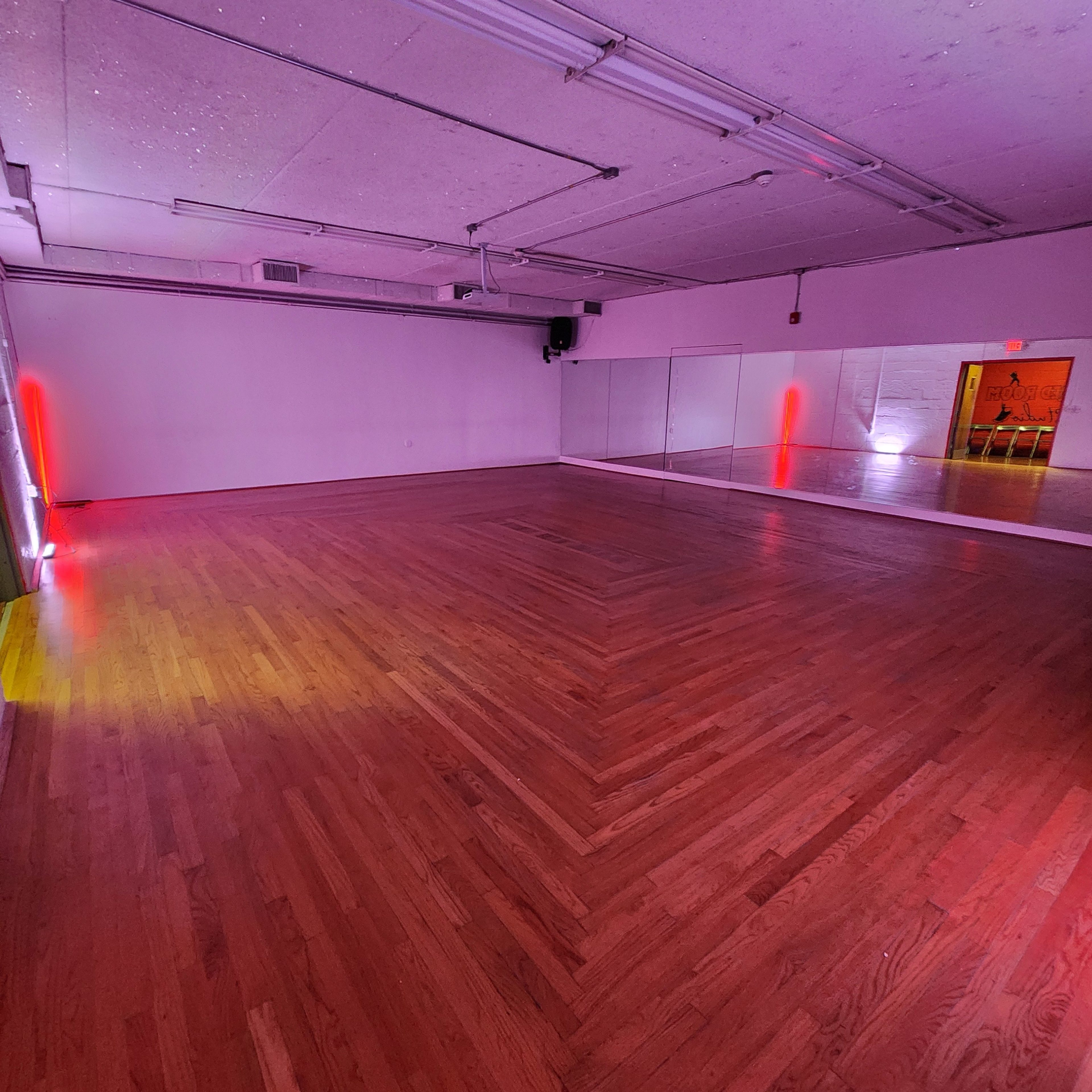 A wooden dance studio with a large mirror on one wall and subtle colored lights illuminating the space.