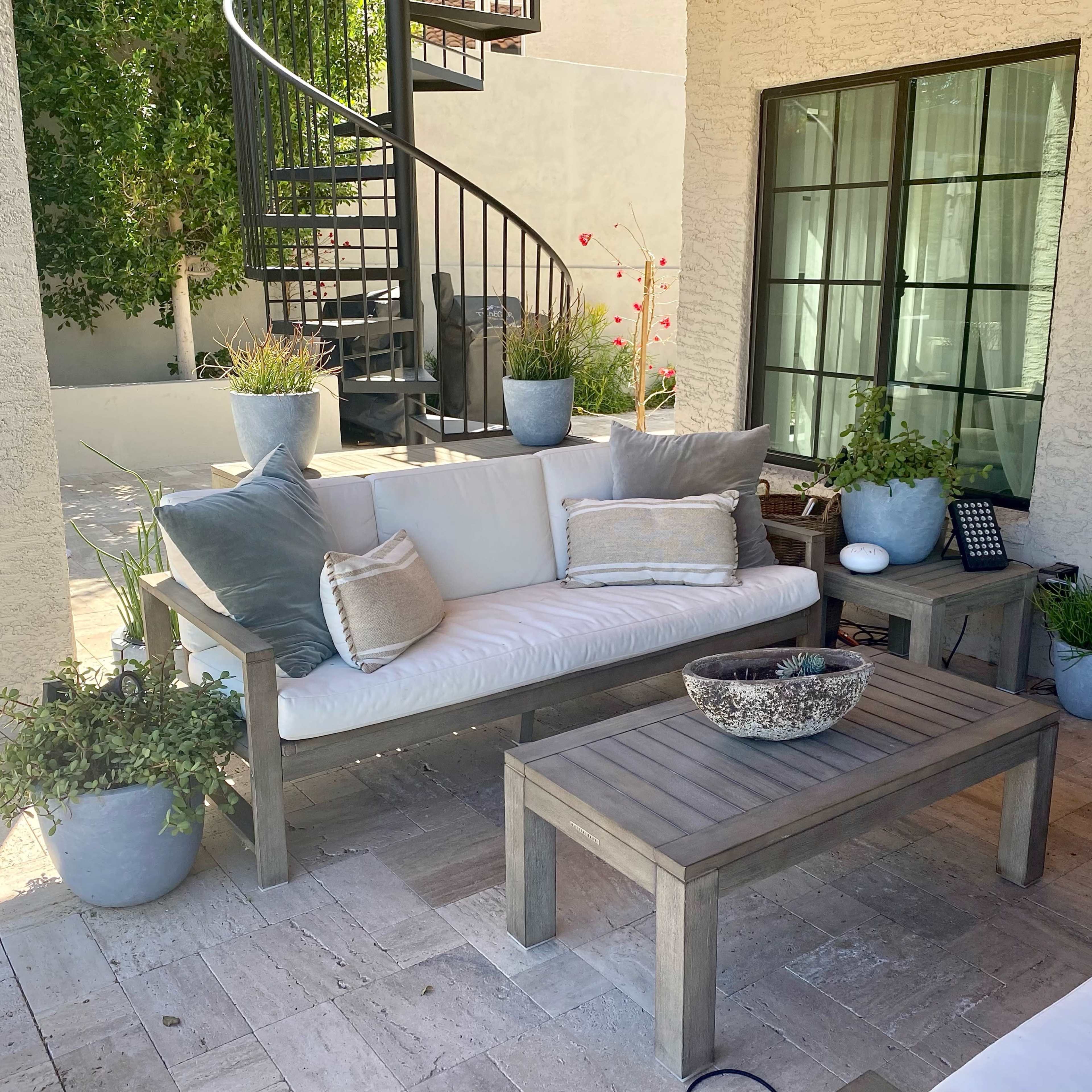 A patio area features a light-colored sofa with cushions, a wooden coffee table, and potted plants arranged around a stone floor.