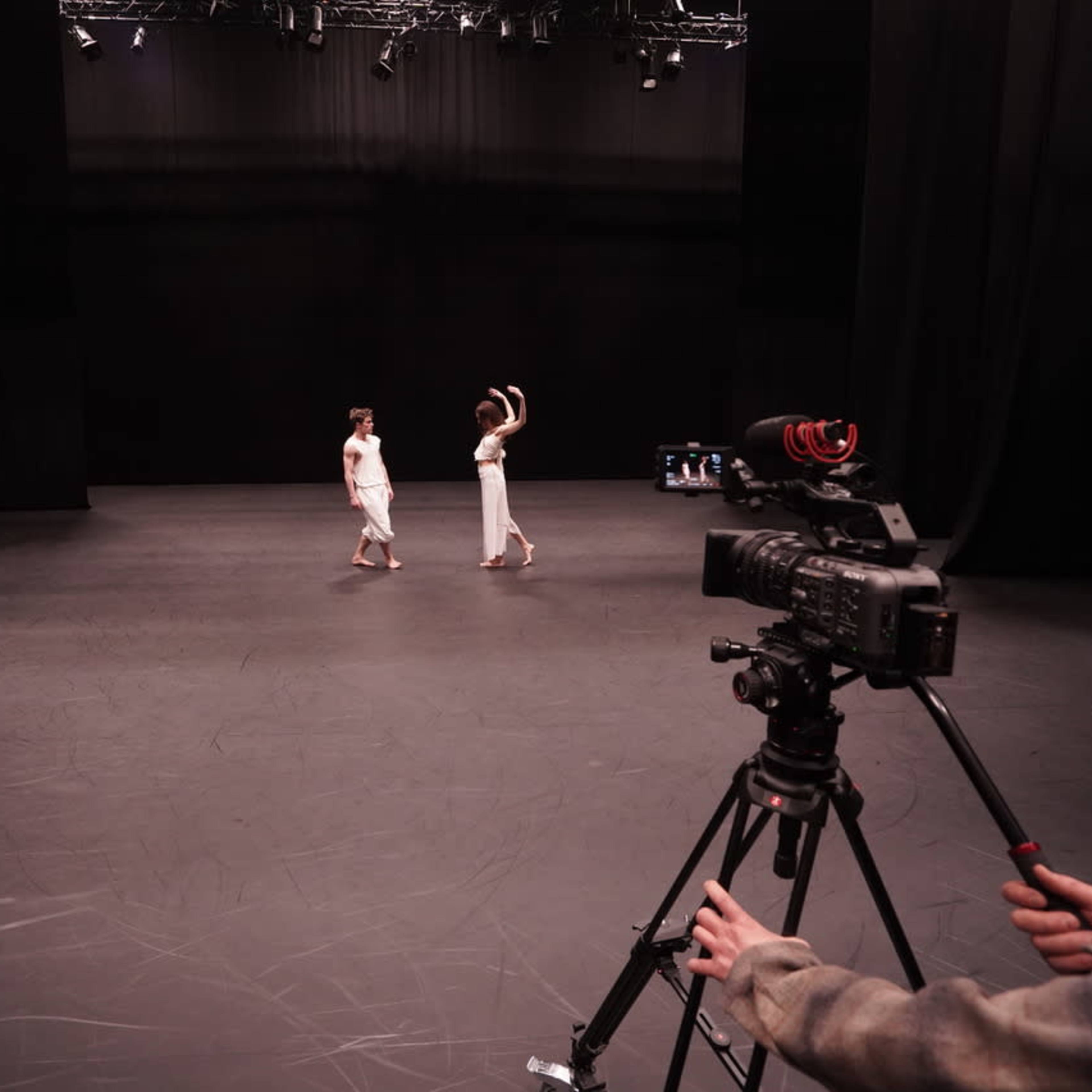 Two dancers are performing on a dark stage while a camera is positioned in the foreground, capturing the scene.