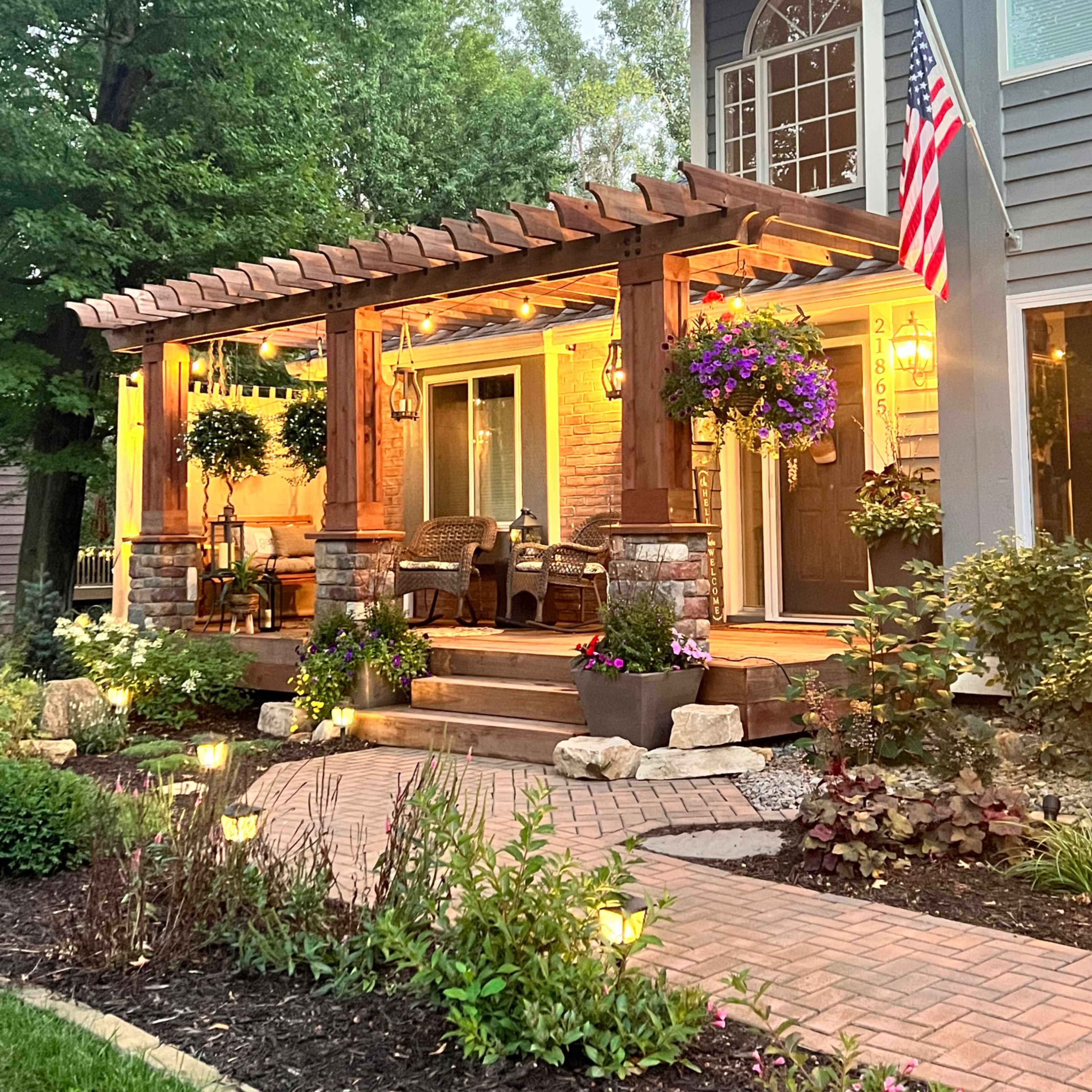 A wooden pergola with hanging baskets and two chairs leads to a well-lit entrance of a two-story home, adorned with American flags and surrounded by landscaped gardens.