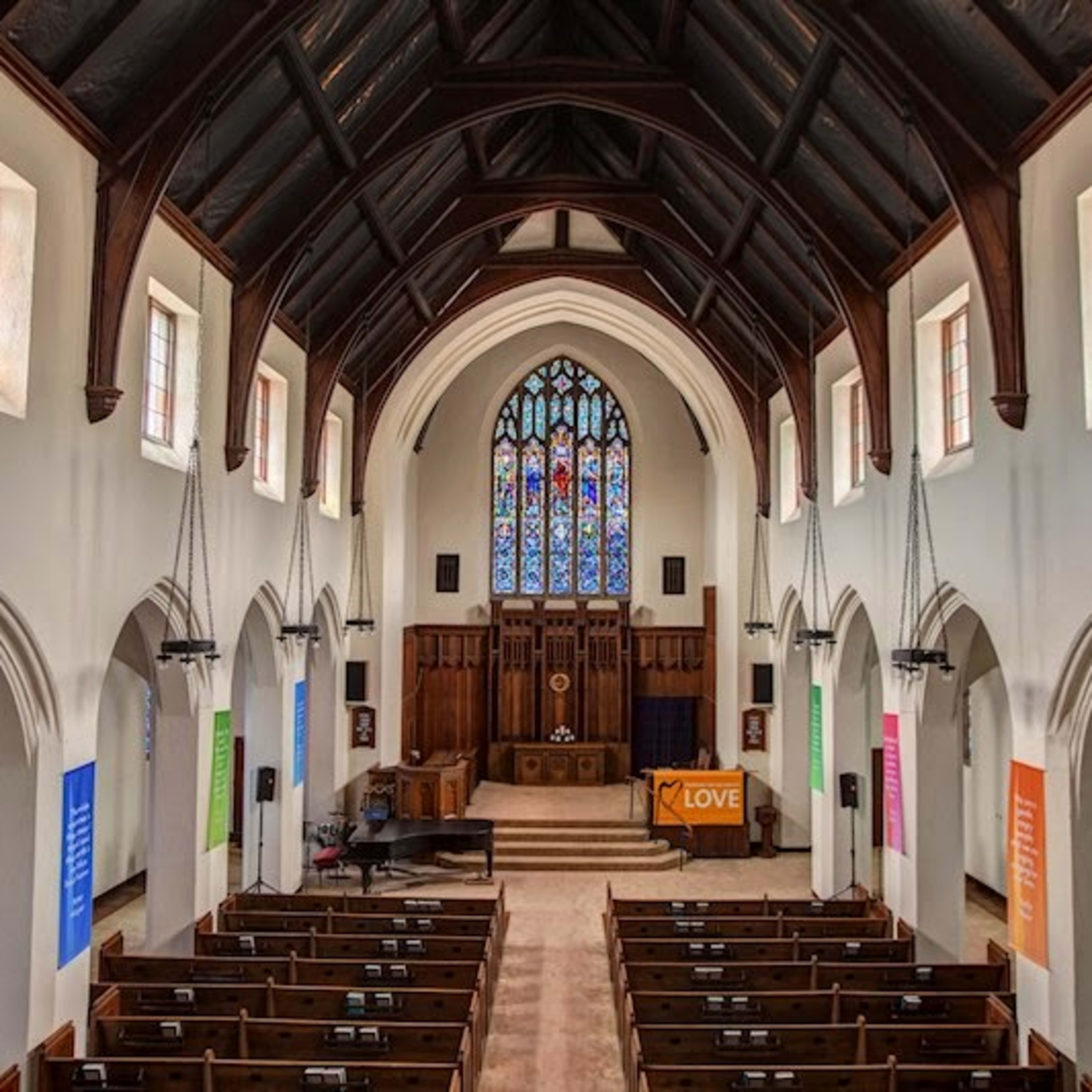 The interior of a church features a high vaulted ceiling, stained glass windows, and wooden benches arranged in rows facing a stage.