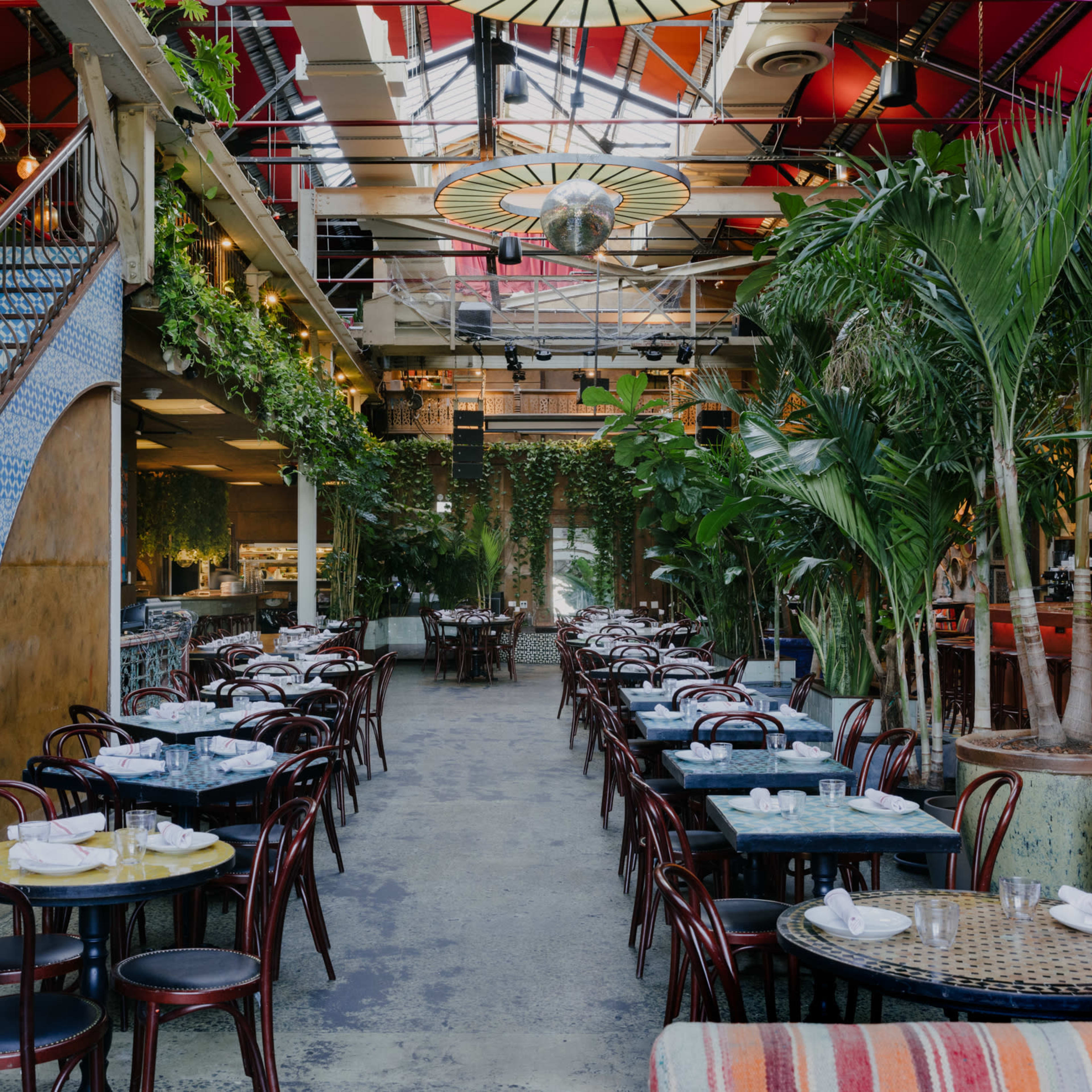 A spacious restaurant interior featuring a spiral staircase, surrounded by greenery and arranged with tables and chairs.
