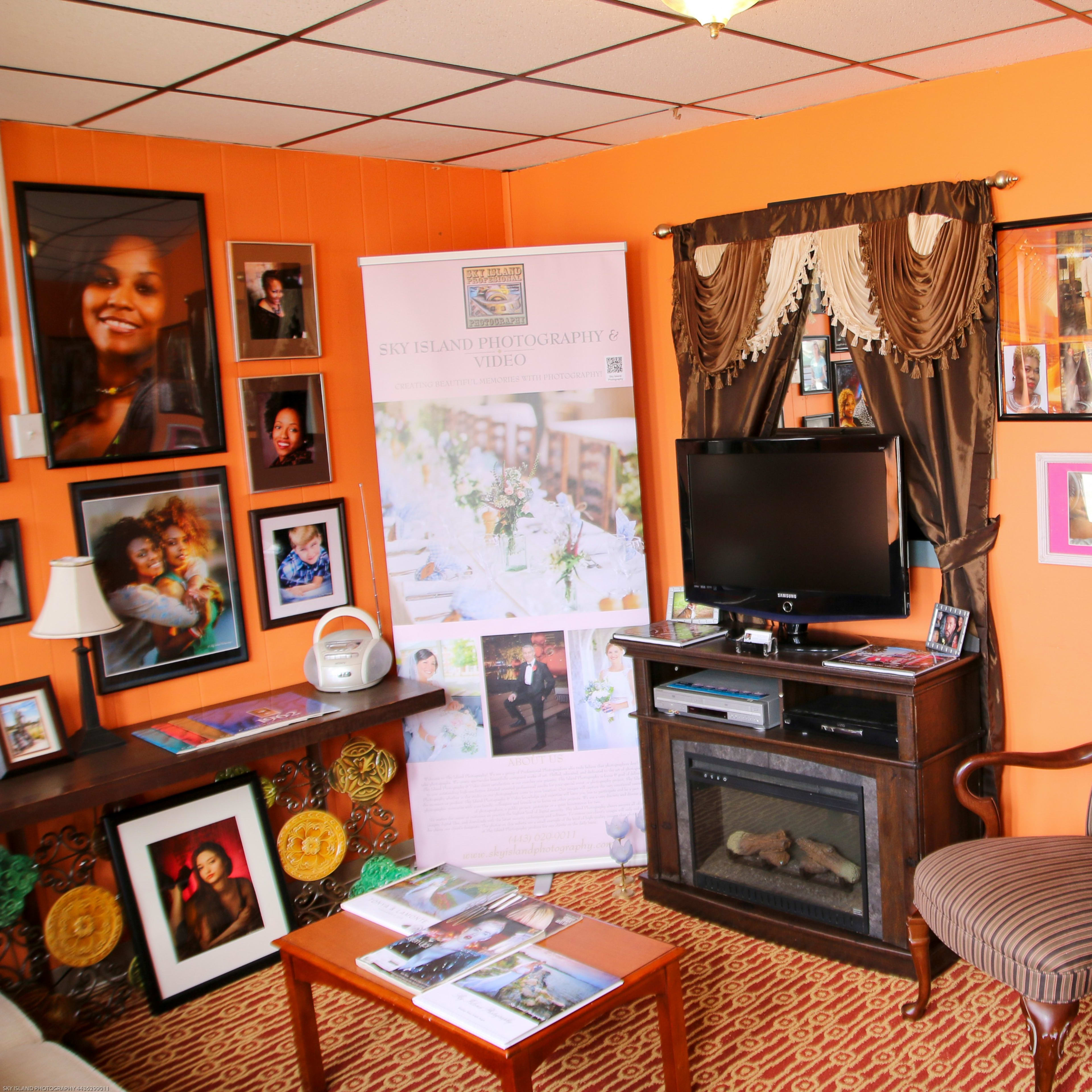 A cozy interior room with orange walls, featuring a television, a decorative chair, framed photographs, and a banner for a photography business.
