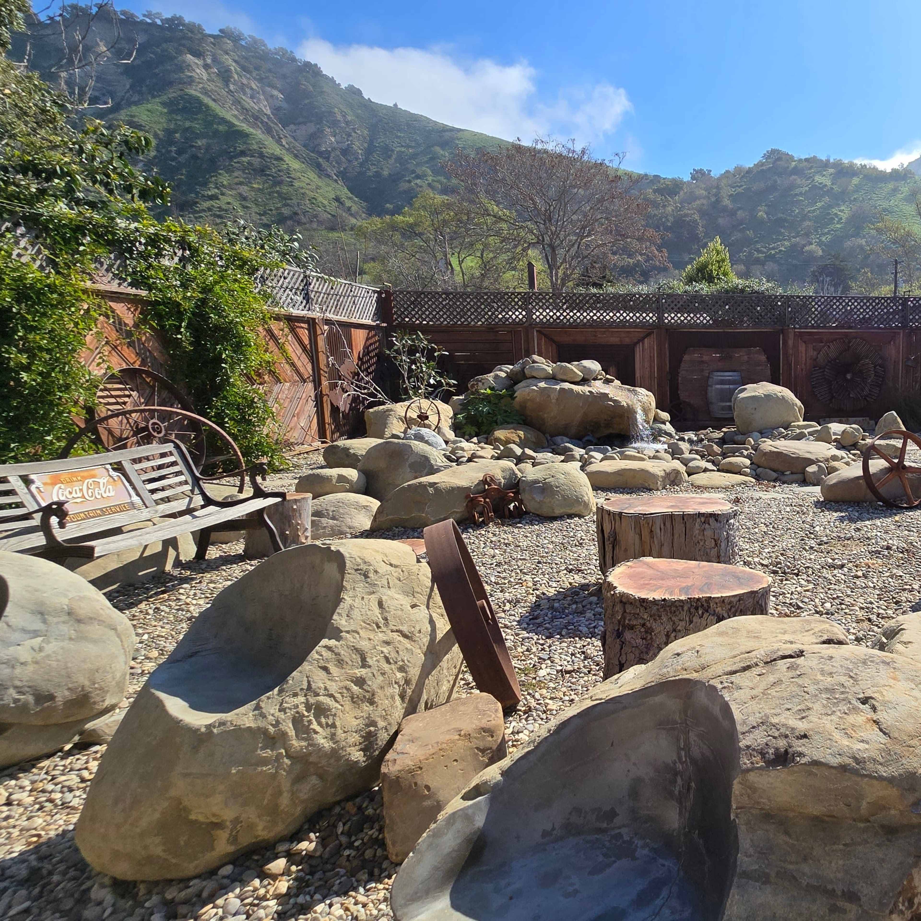 A rock garden with large stones, wooden logs, and a bench, surrounded by a wooden fence and greenery under a clear blue sky.