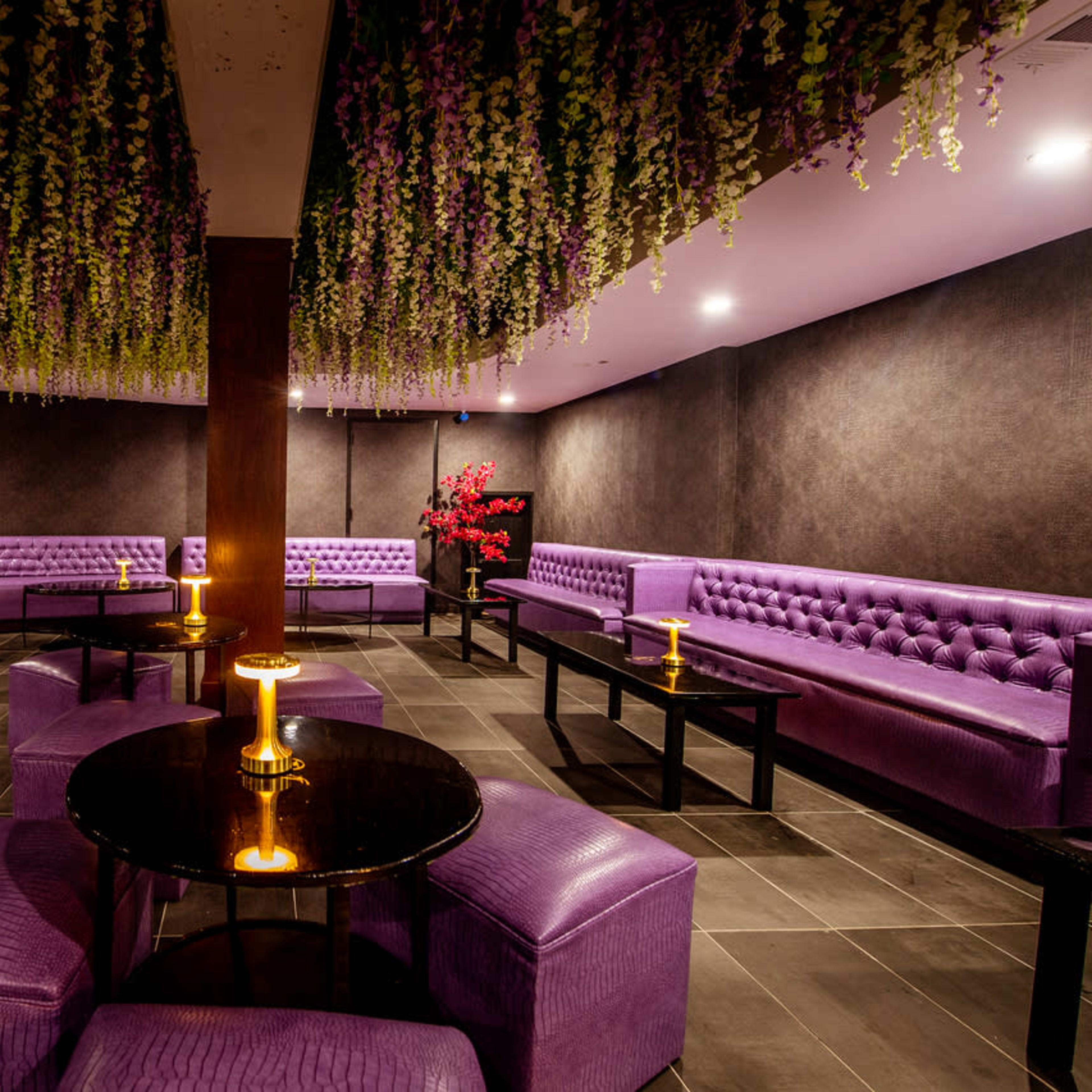 The scene features a dimly lit lounge with purple tufted seating, black tables, and decorative floral arrangements hanging from the ceiling.