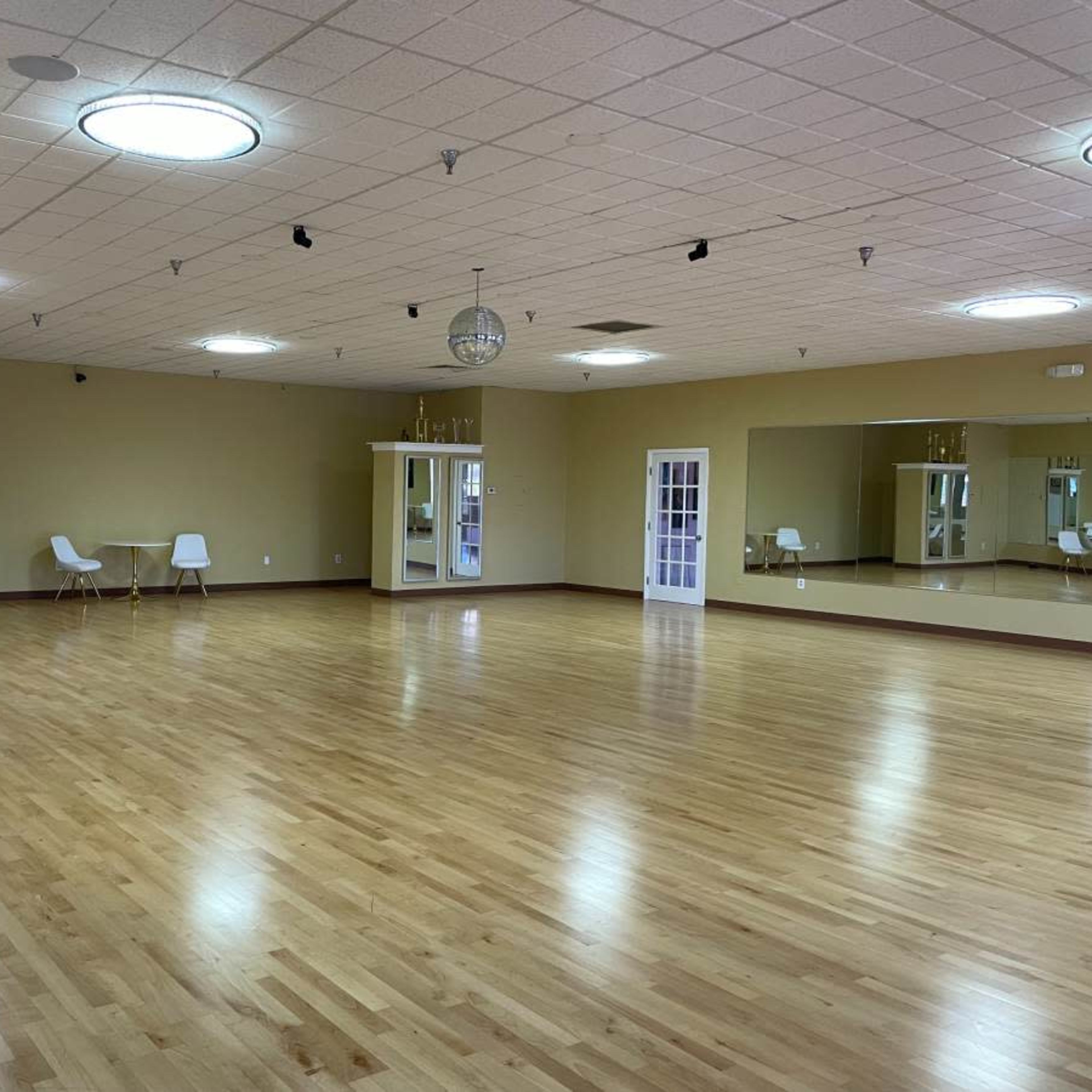 A spacious dance studio with wooden floors, mirrored walls, and a few mirrors and trophies displayed along one side.