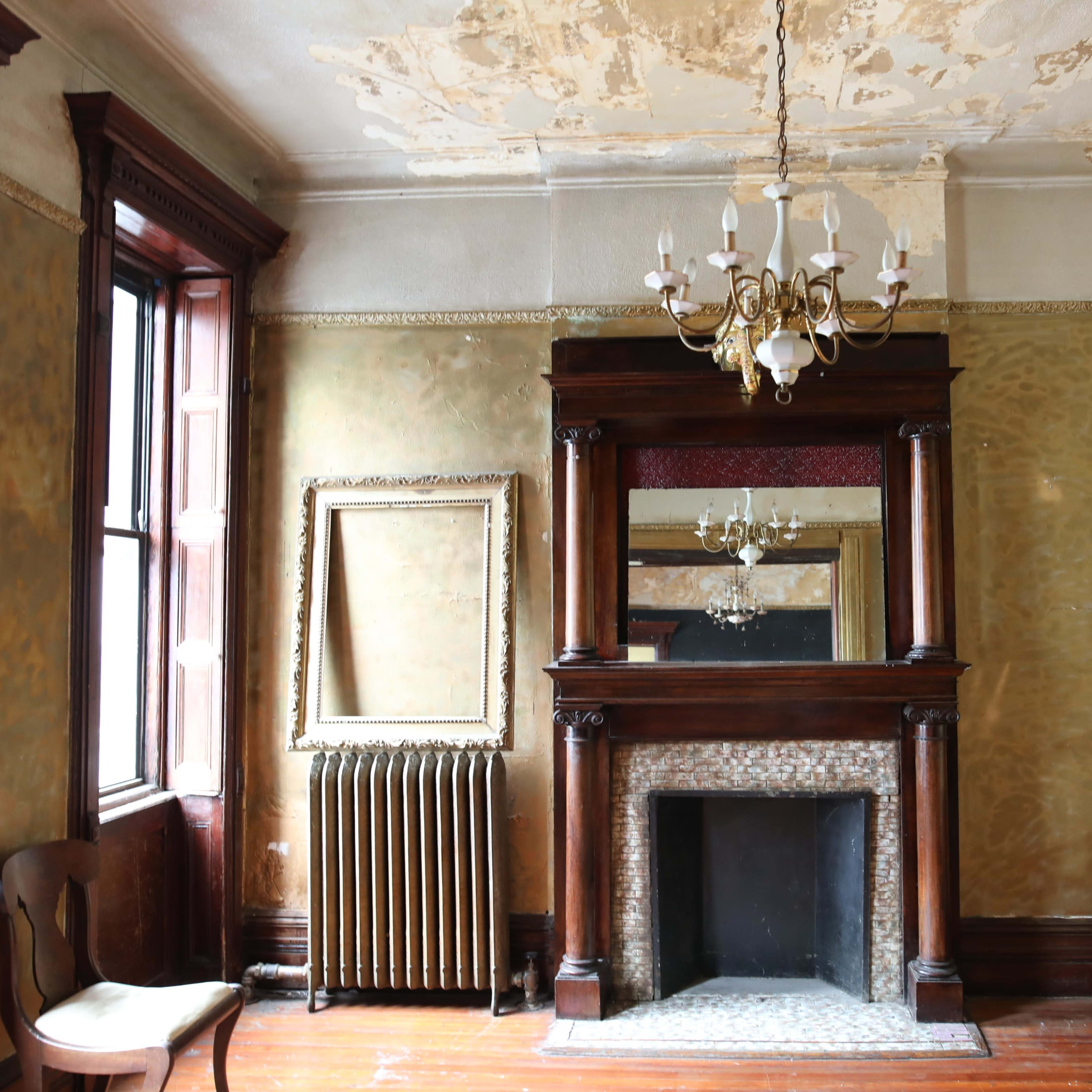 The room features a wooden fireplace with a mirror above it, a vintage chandelier, and large windows alongside a battered wall and faded wallpaper.
