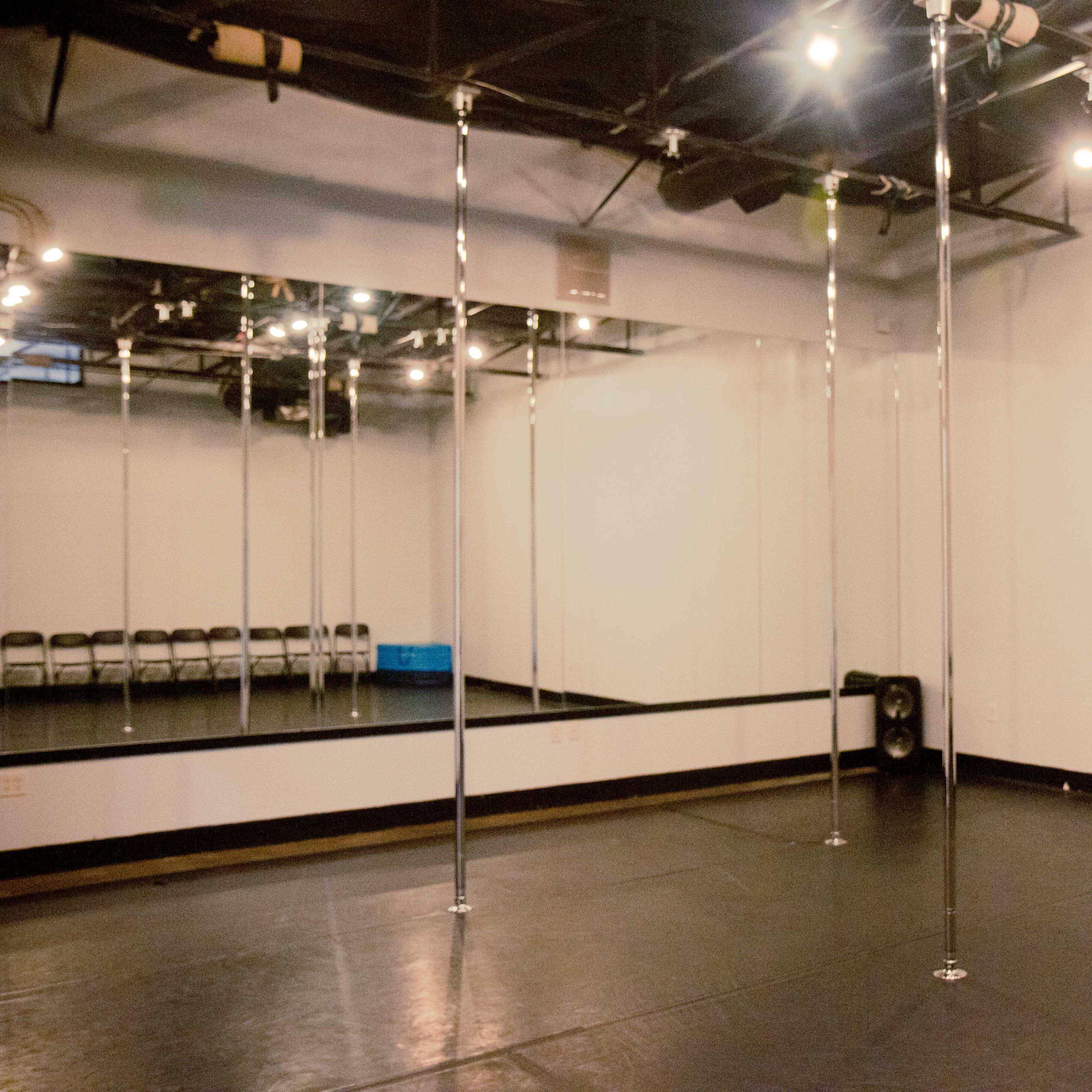 A studio with several polished poles, a large mirror, and a black floor, illuminated by overhead lights.
