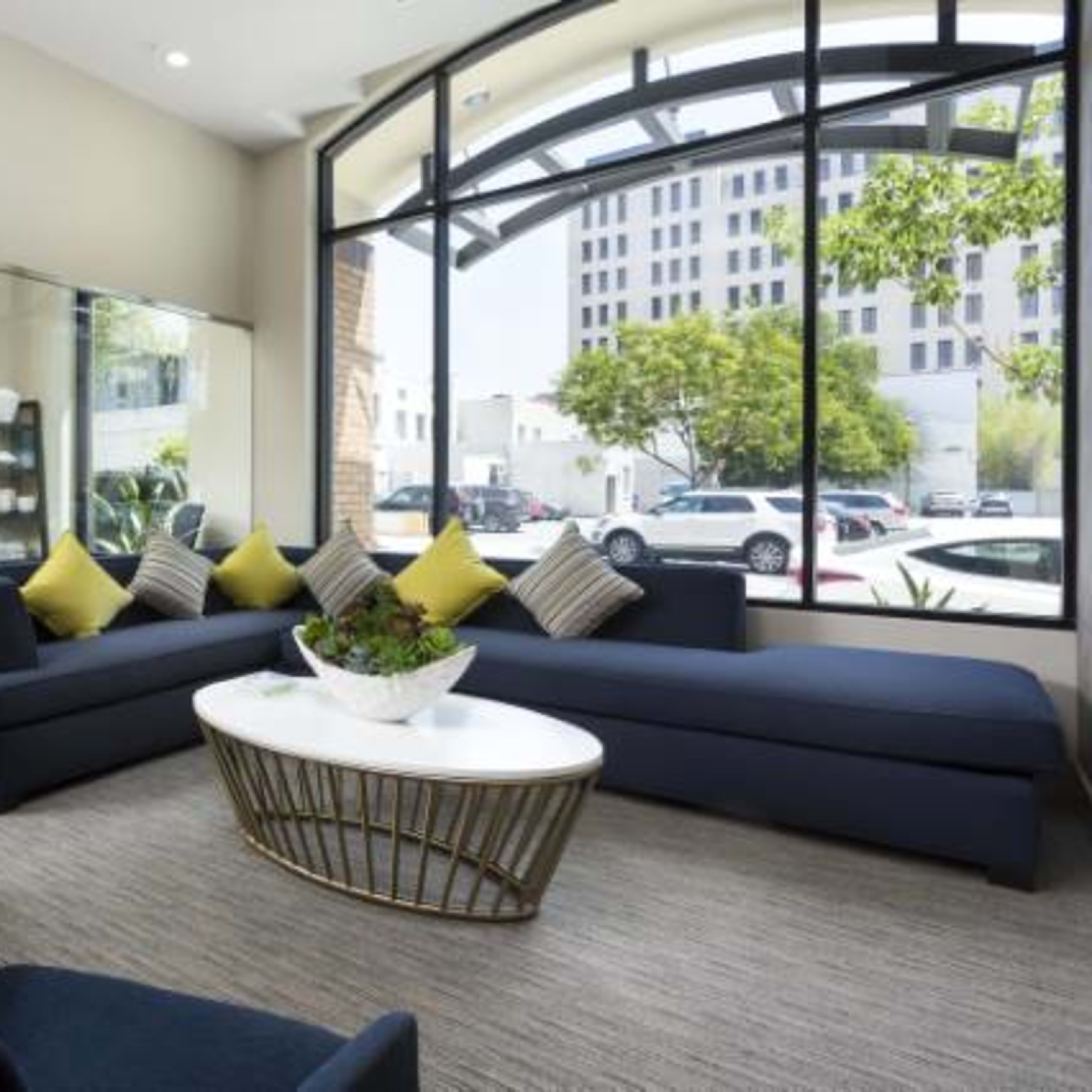 A modern lounge area features a navy sectional sofa, a round coffee table, and large windows overlooking a city street.