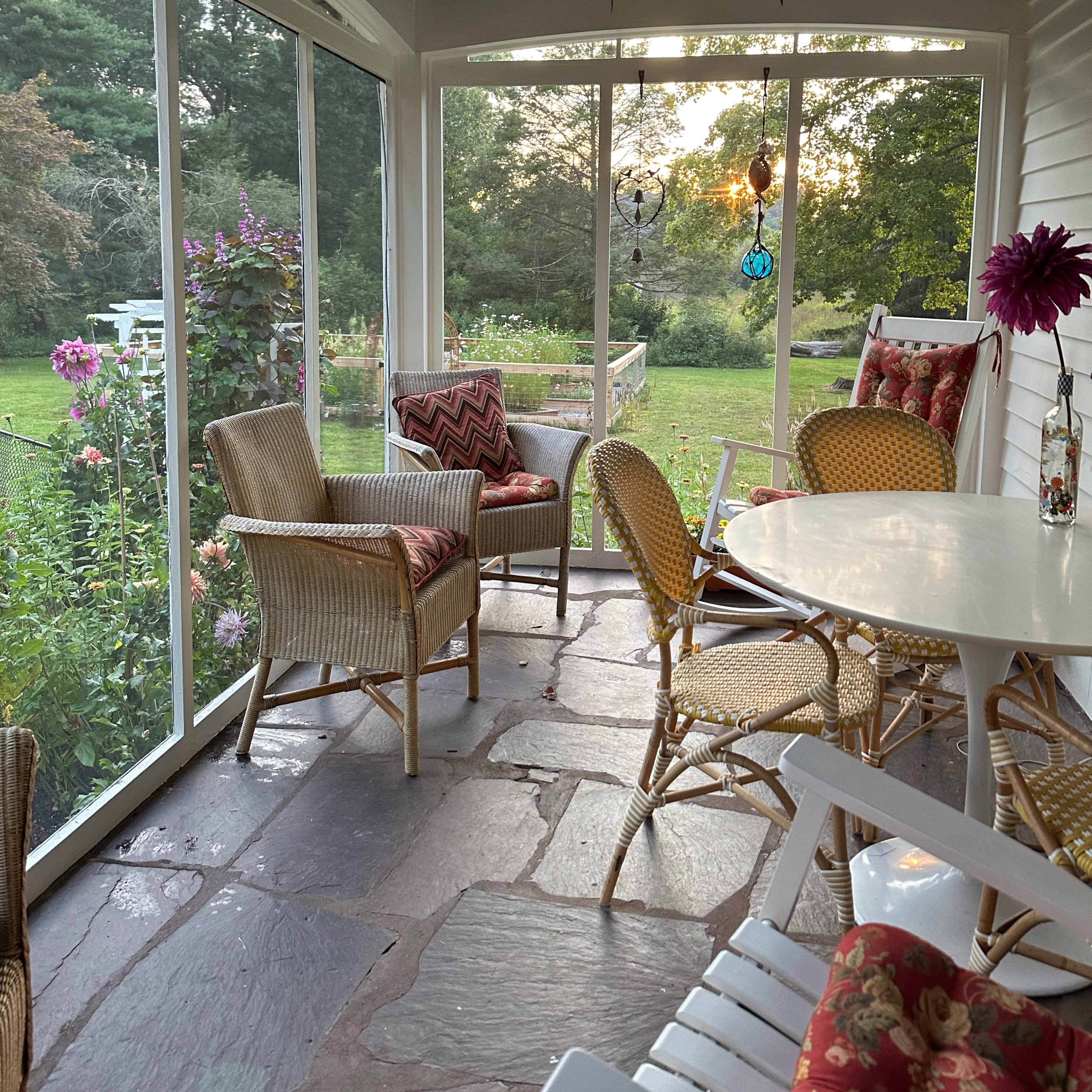 A screened porch with wicker chairs and a small table, surrounded by greenery.