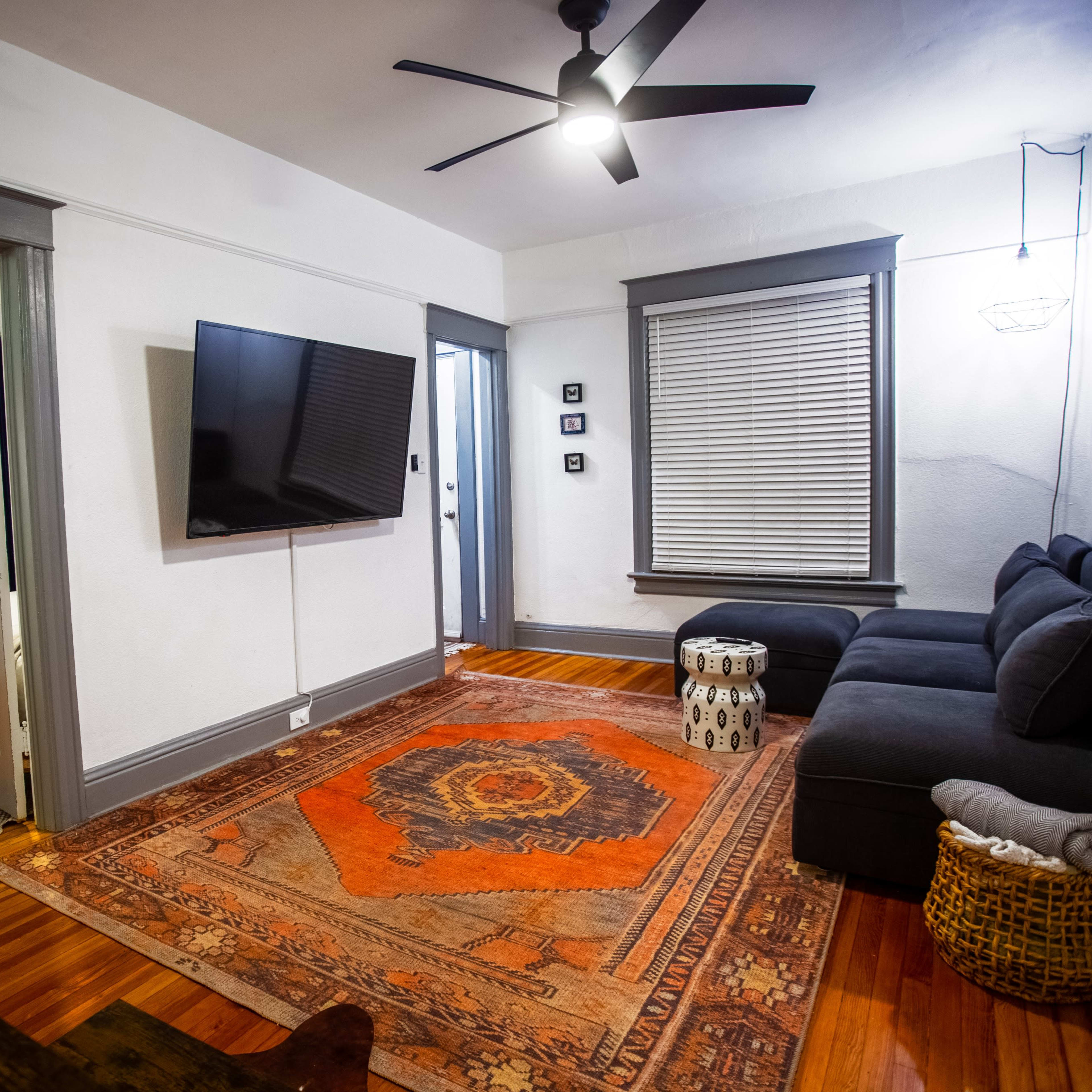 A living room with a dark couch, a large television mounted on the wall, and a colorful area rug on wooden flooring, leading to a doorway with a bed visible in the adjacent room.