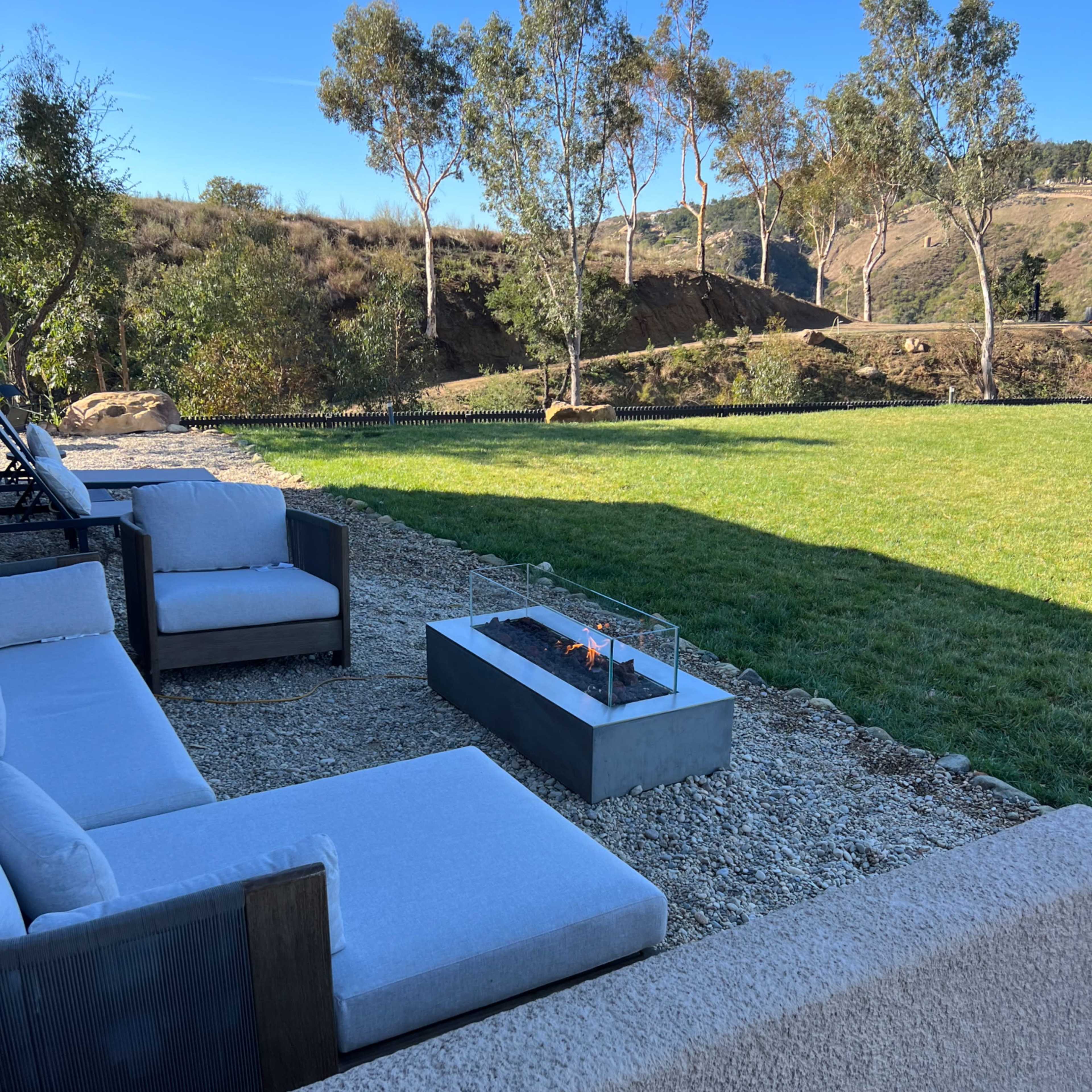 A modern outdoor seating area features plush sofas arranged around a fire pit, with a backdrop of rolling hills and greenery.