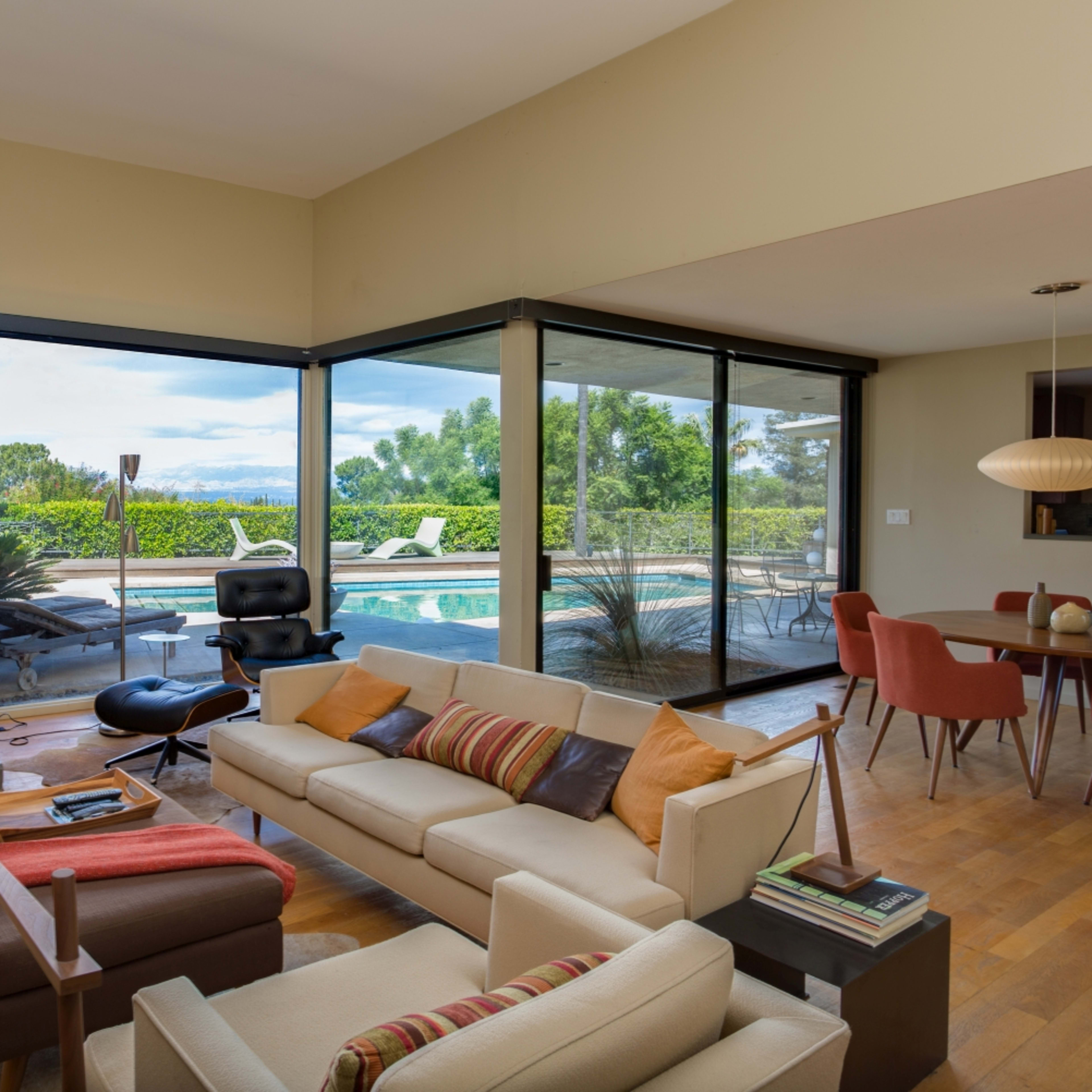 A modern living room features a large glass window overlooking a pool area and lush greenery, with a neutral-colored sofa and a simple dining area.