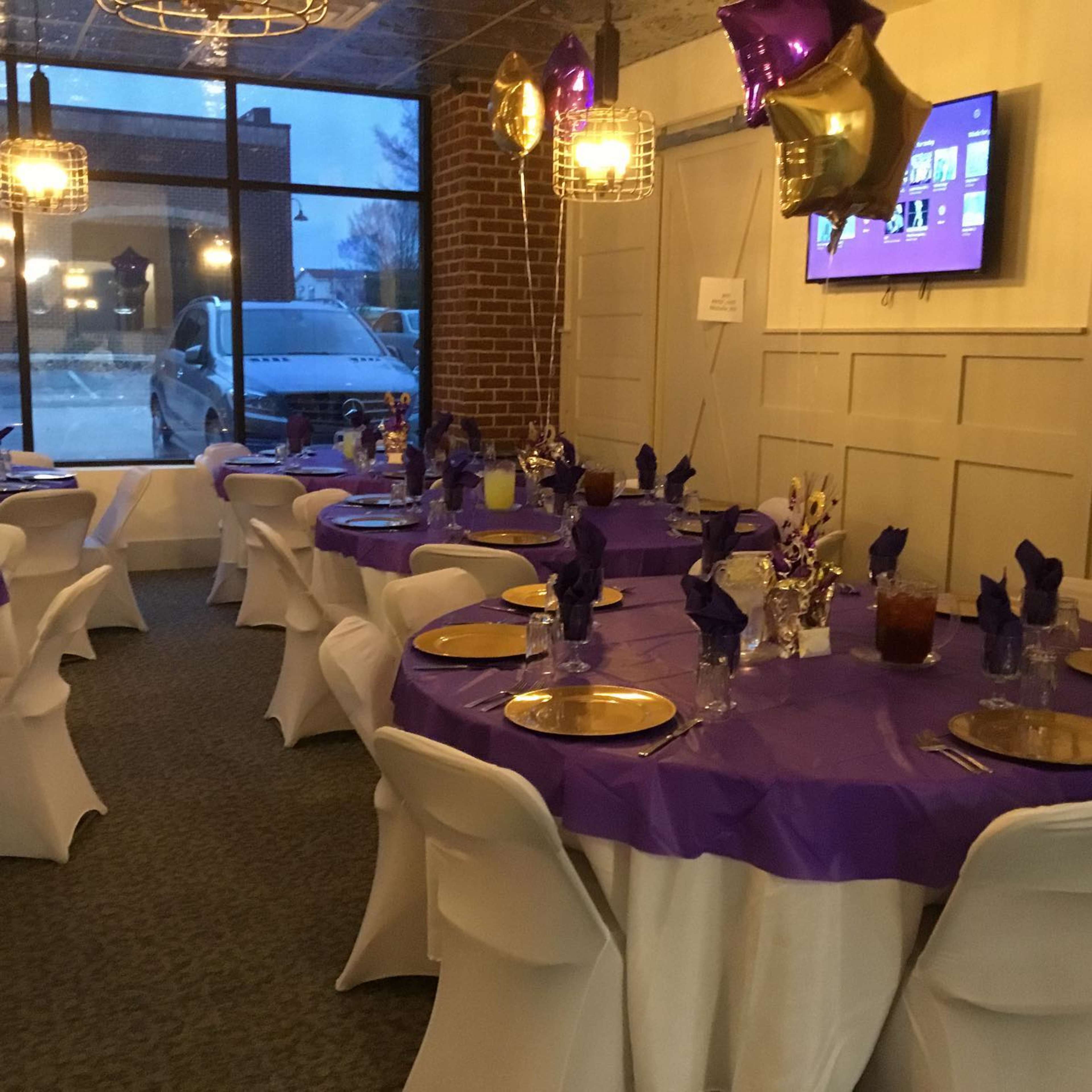 A decorated dining area with round tables covered in purple tablecloths, each set with plates and glasses, and a television mounted on the wall displaying a menu or event information.