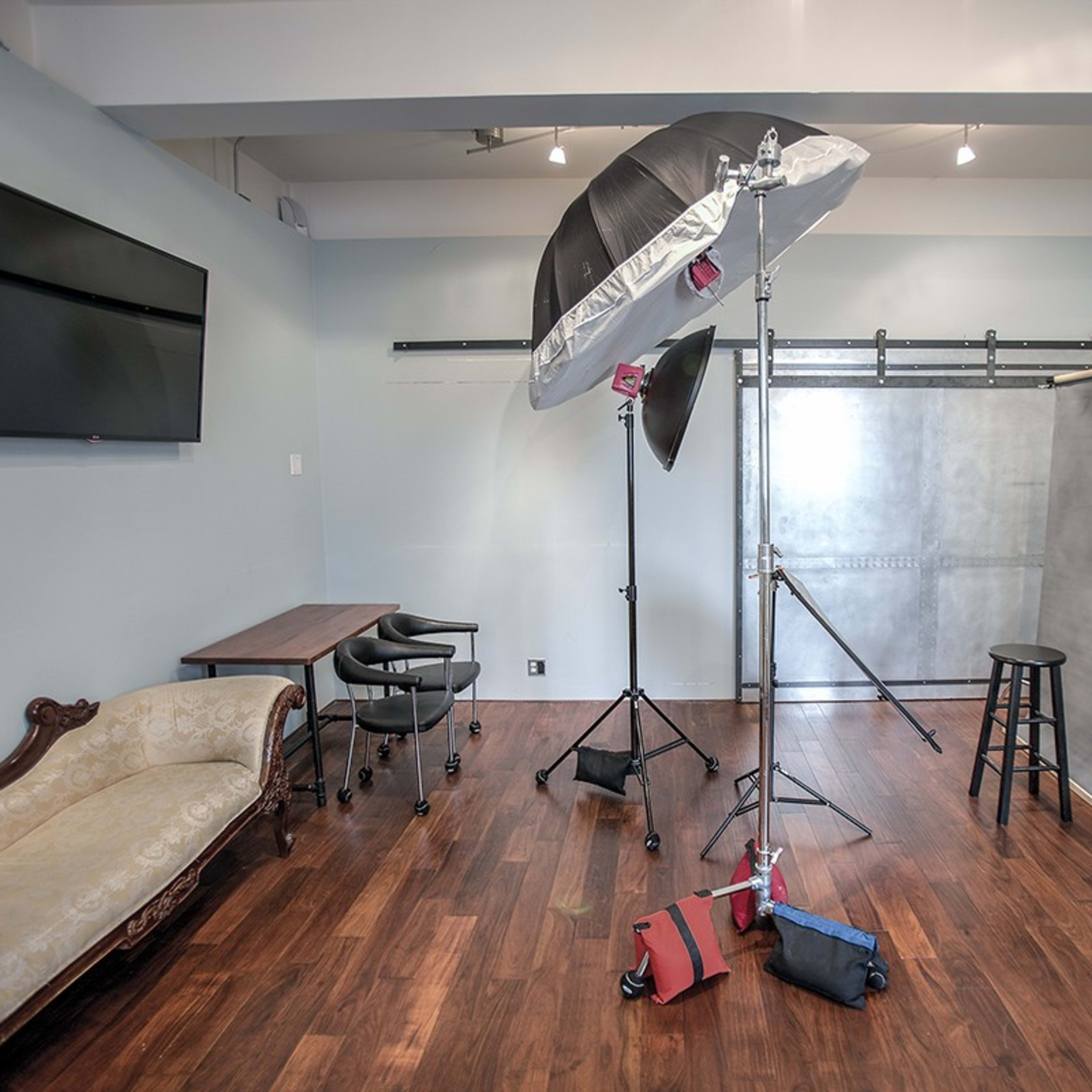 A photography studio featuring a lighting setup, a backdrop, a small table with chairs, and a chaise lounge against the wall.