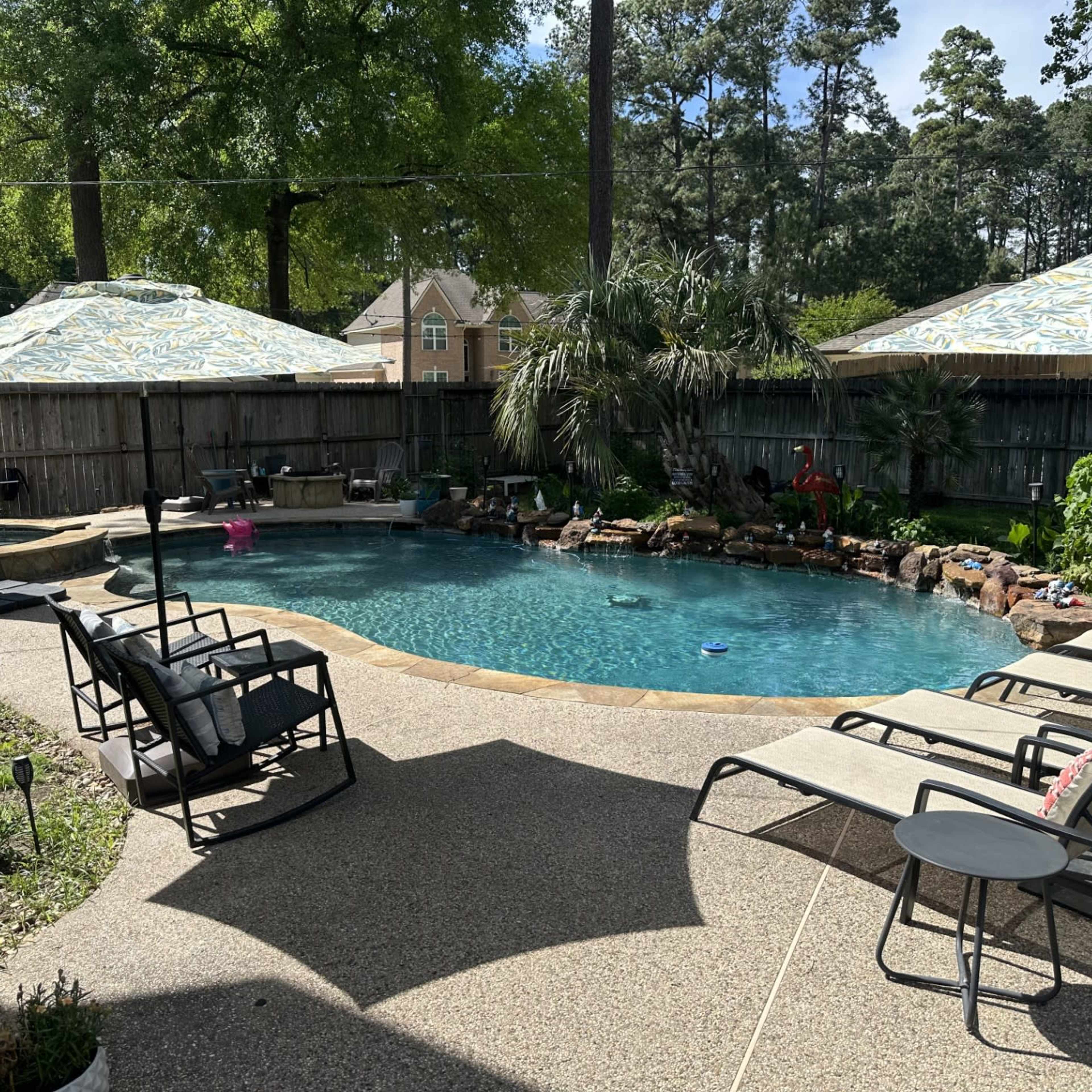 A backyard pool area features a clear blue swimming pool surrounded by lounge chairs and shaded by umbrellas.