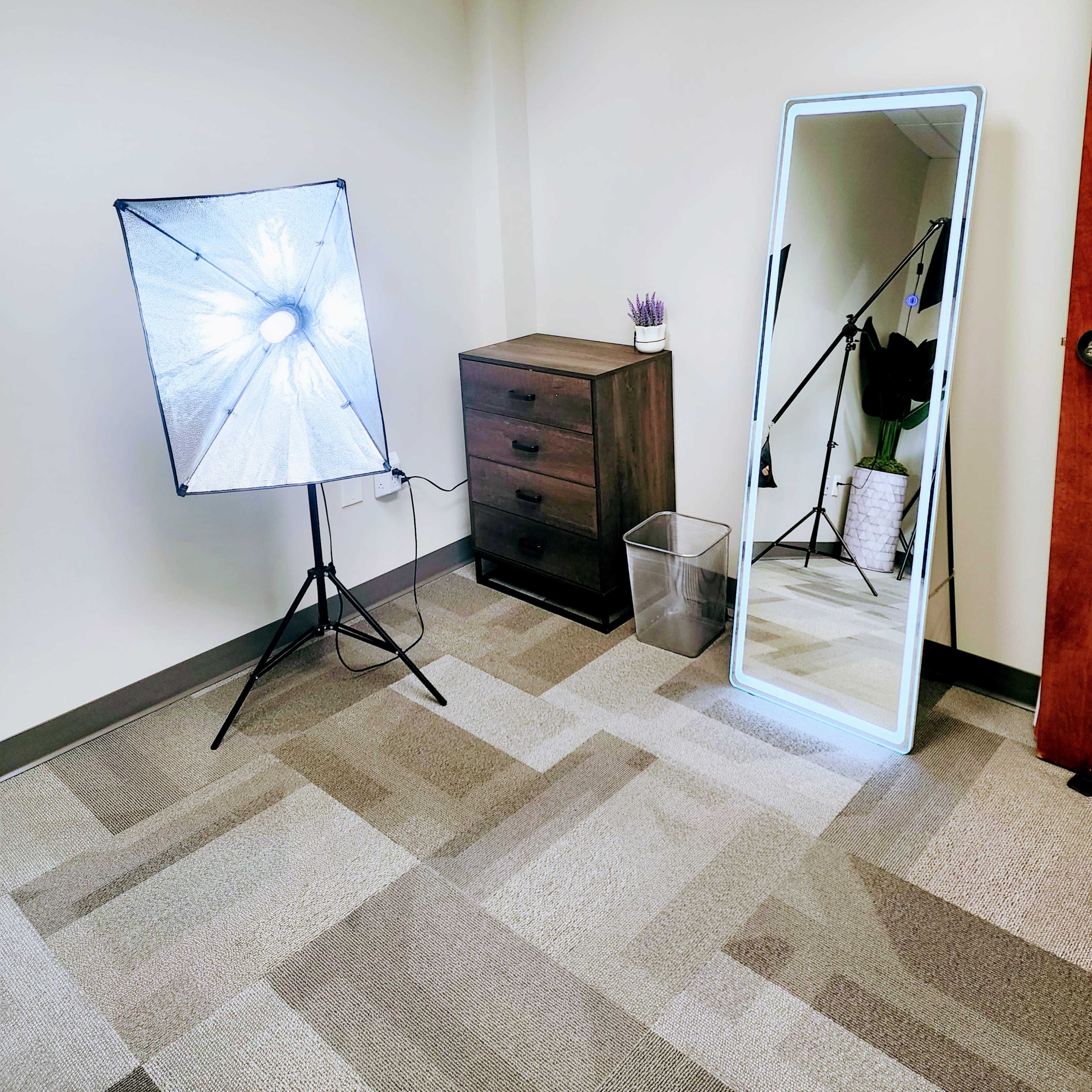 A corner of a room featuring a softbox light, a tall mirror, a small dresser with drawers, and a wastebasket.