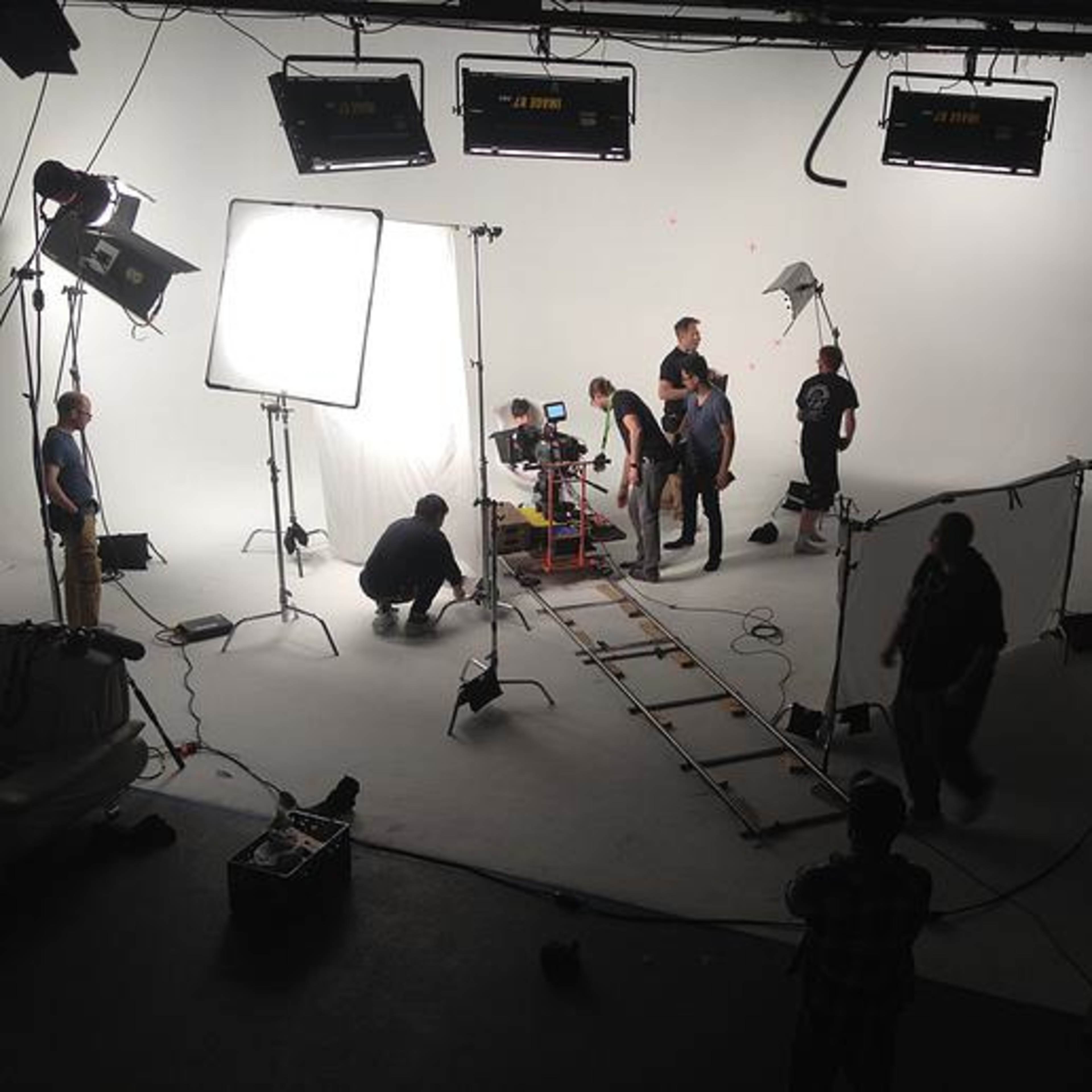 A film crew sets up camera equipment on a studio set with a white backdrop and softbox lights.