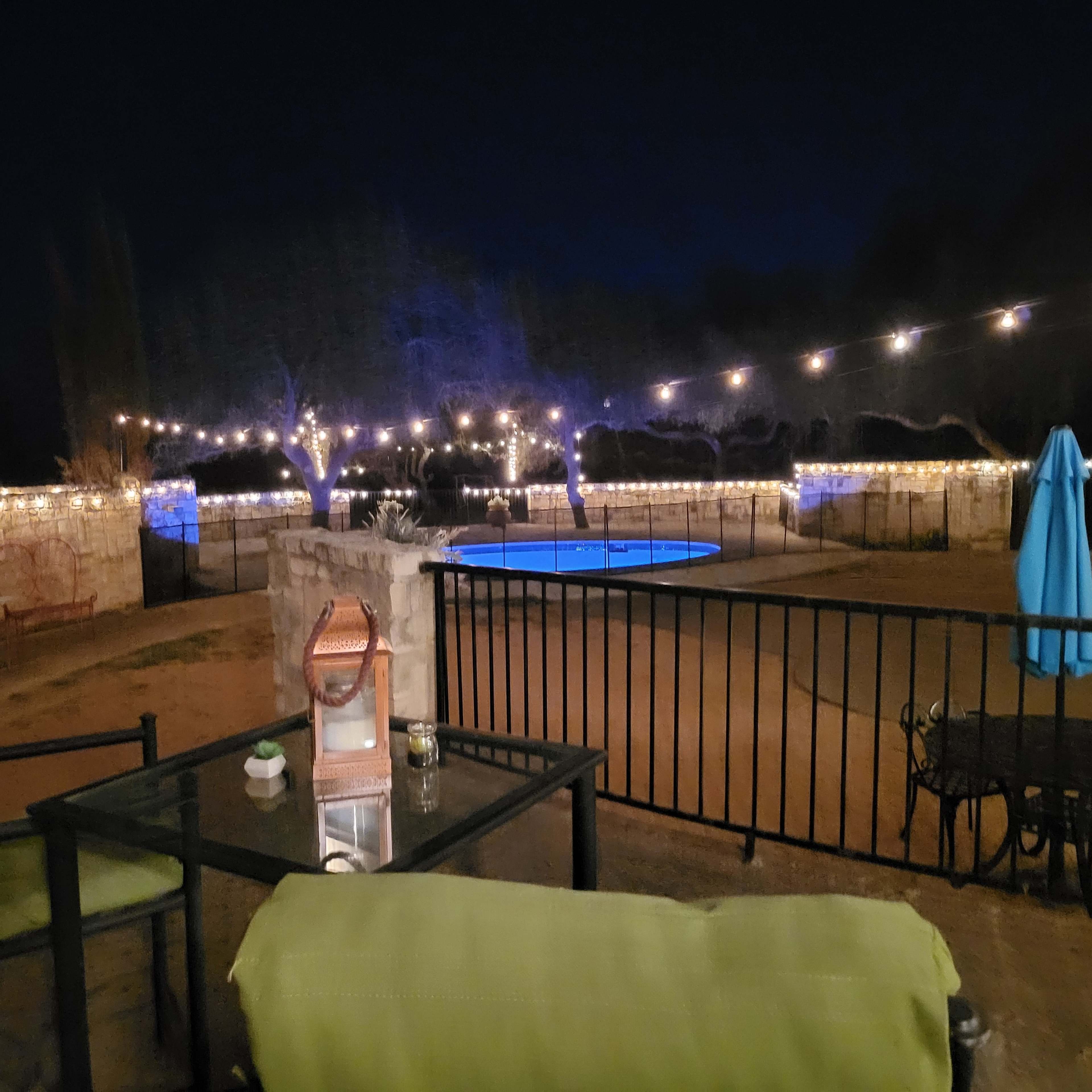 A lounge area with green chairs overlooks a pool illuminated by string lights at night.